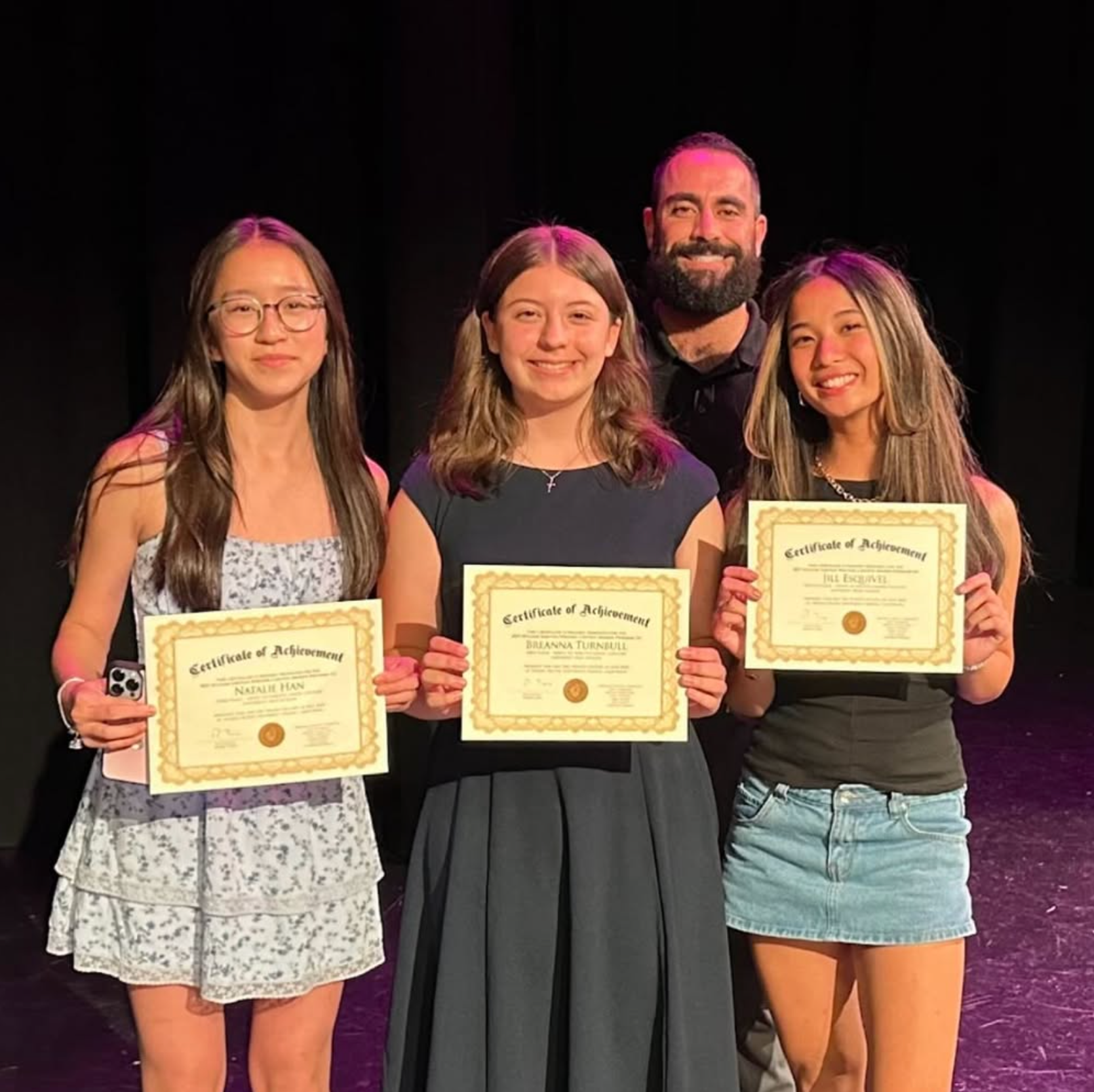 Three young women and an adult man standing on a stage, holding certificates of achievement and smiling for the camera.