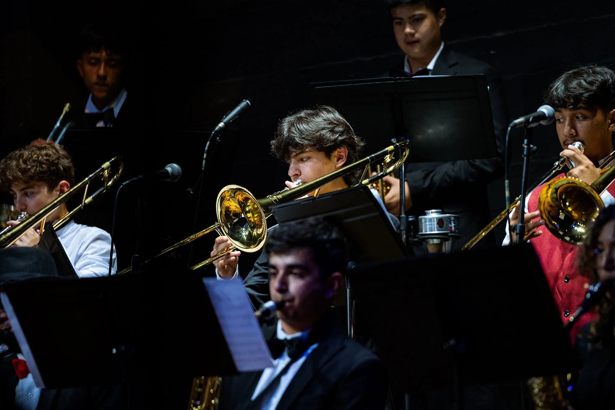 A jazz band performing on stage with several young musicians playing brass instruments such as trombones, wearing formal attire, in a dimly lit room.