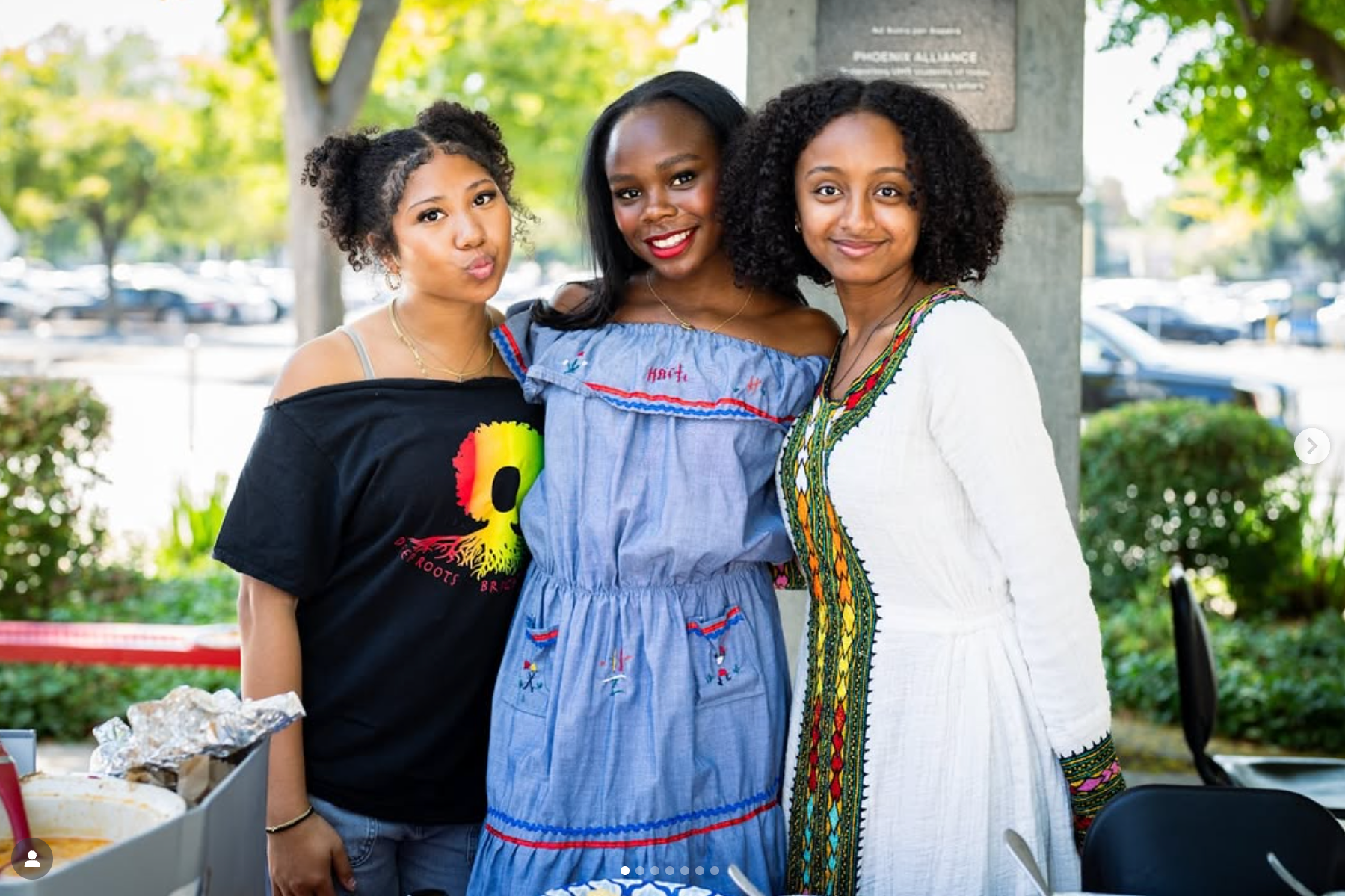 Three women standing outdoors side by side, smiling at the camera. Two women are on either side of a woman in a blue dress. The background shows trees and parked cars.