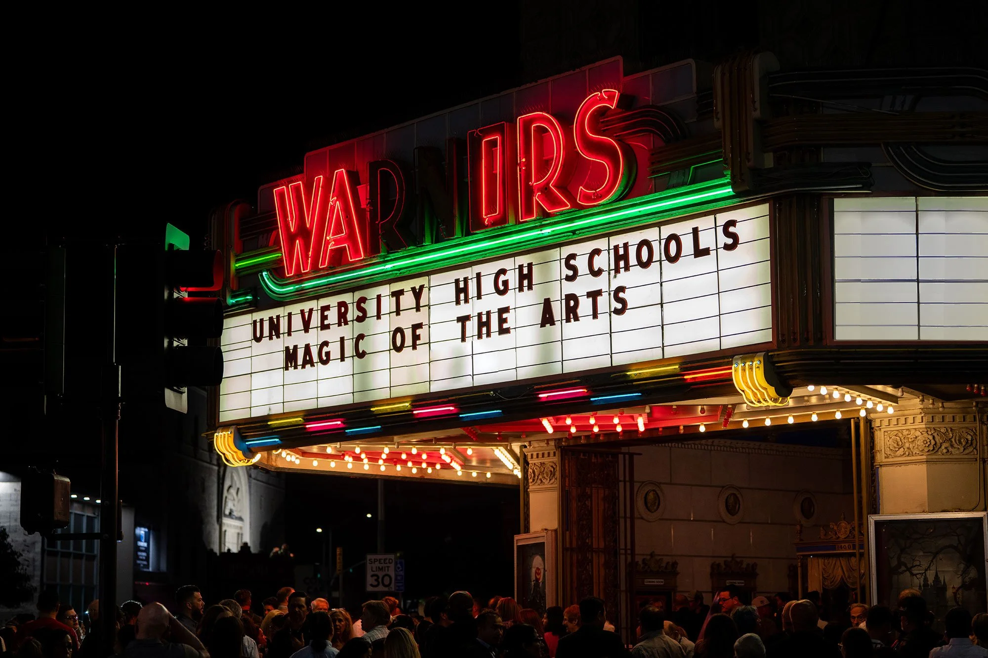 Night shot of an illuminated theater marquee with neon signs reading 'WARNERS' and a message board listing 'UNIVERSITY HIGH SCHOOLS' and 'MAGIC OF THE ARTS'. Crowd gathered outside.