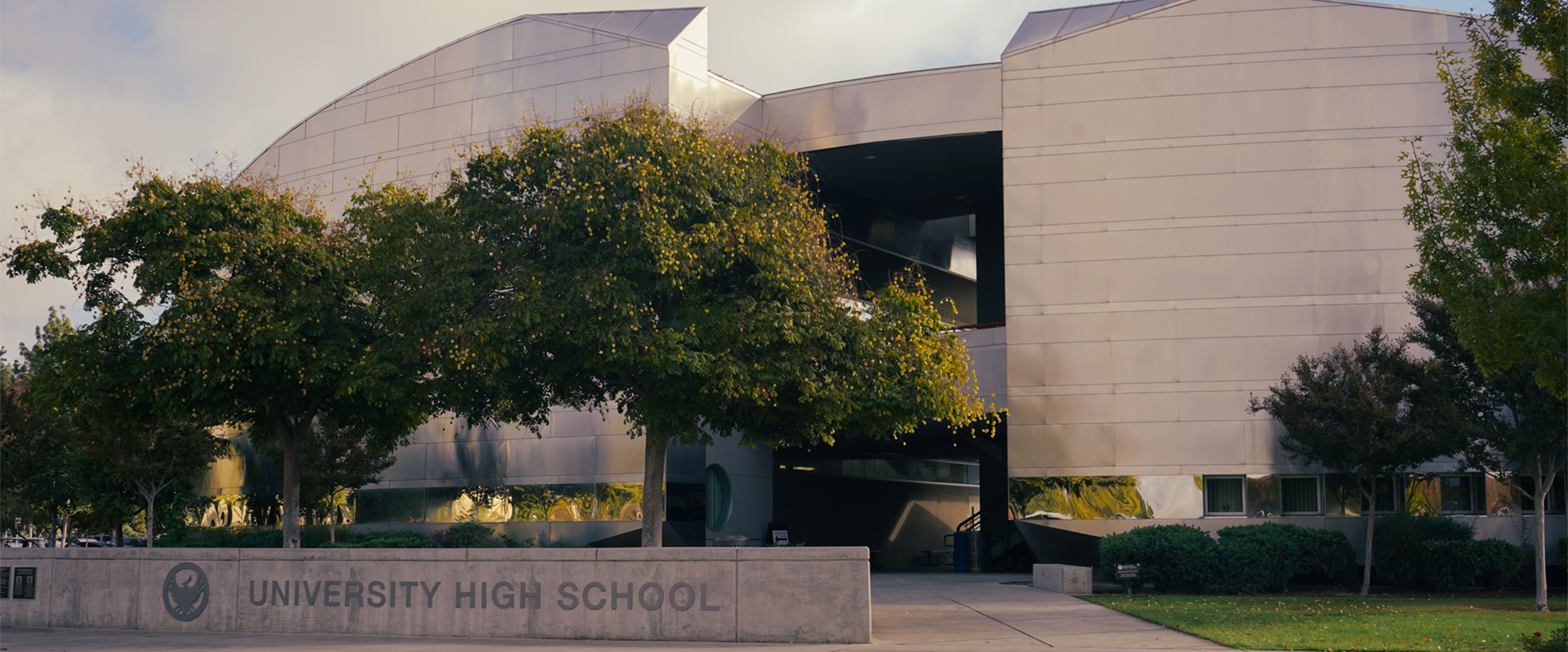 The University High School building with a modern, curved metallic exterior, surrounded by trees and a landscaped area. The school's name is on a concrete sign in the foreground.