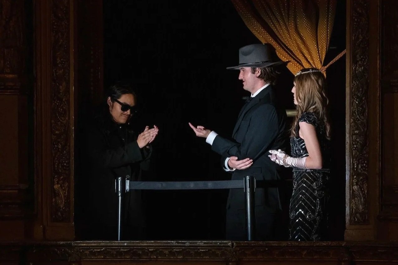A man in a suit and hat stands behind a waist-high barrier, holding out his hand. A woman in a black dress with gloves stands behind him. A man with sunglasses and black clothing is in front of them, with hands clasped, facing the man with the hat. They appear to be on a stage or in a theatrical setting with ornate wooden framing and heavy curtains.