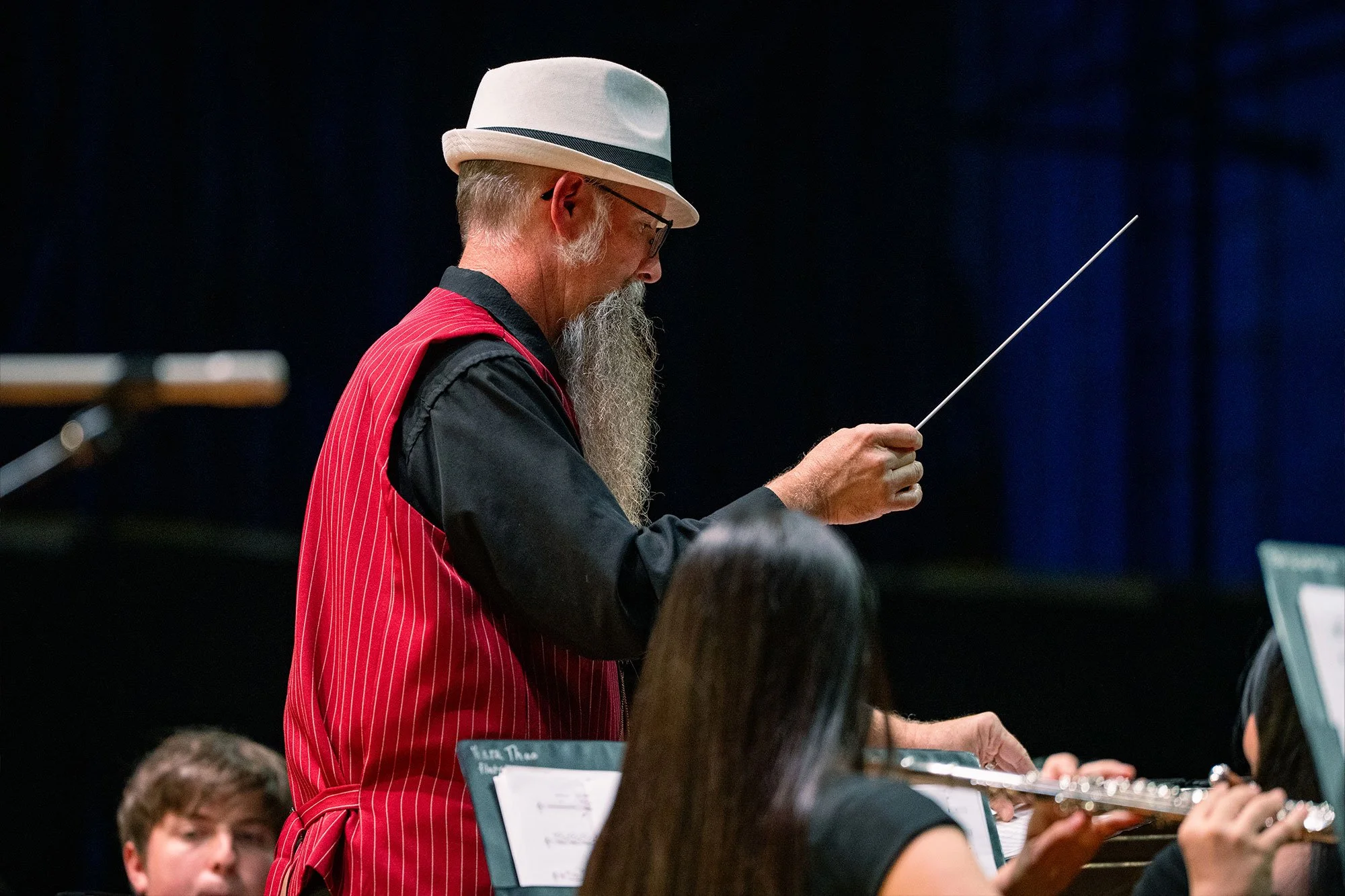 An older man with a long beard and glasses, wearing a white fedora, a red vest over a black shirt, conducting an orchestra with a baton.