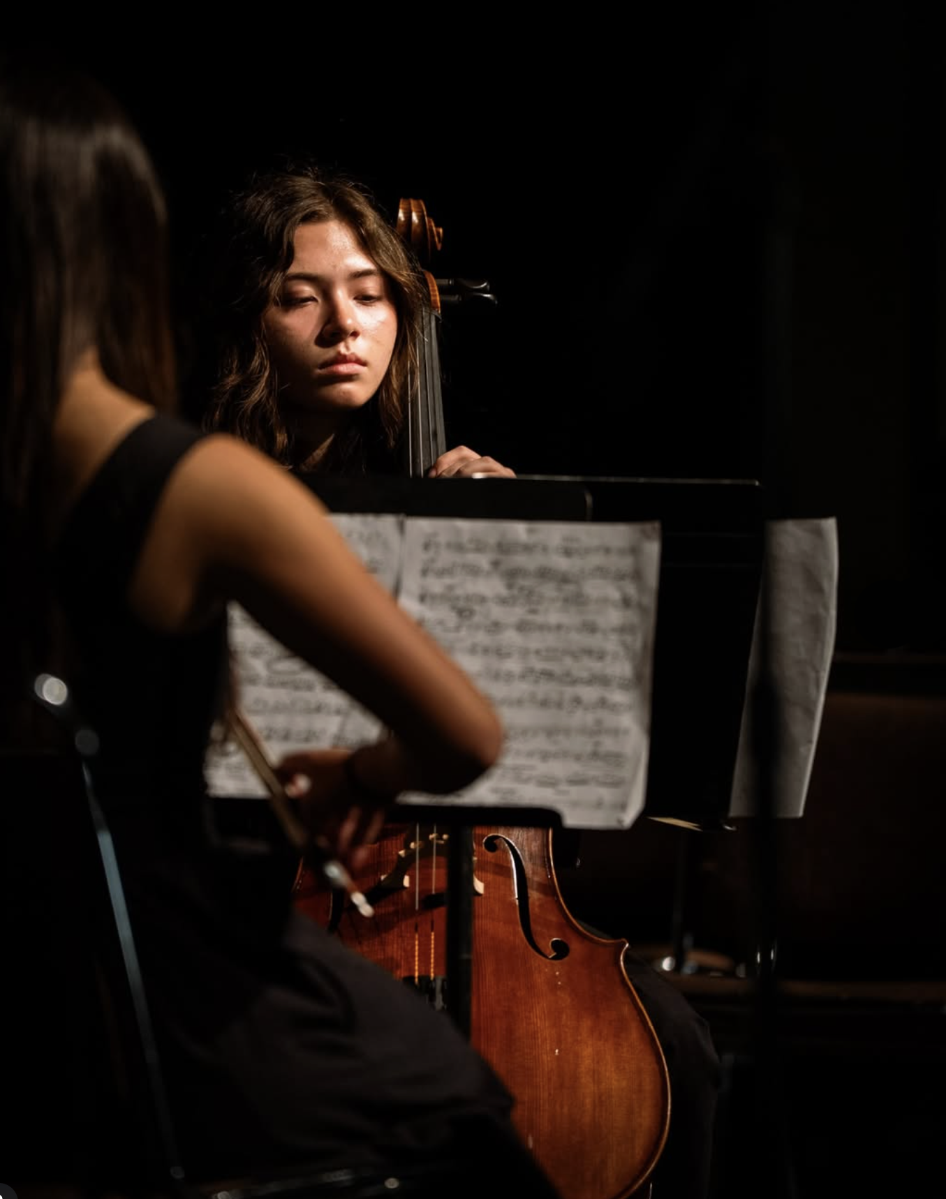 Two young women playing string instruments, one on piano and the other on cello, in a darkened music studio or stage setting.