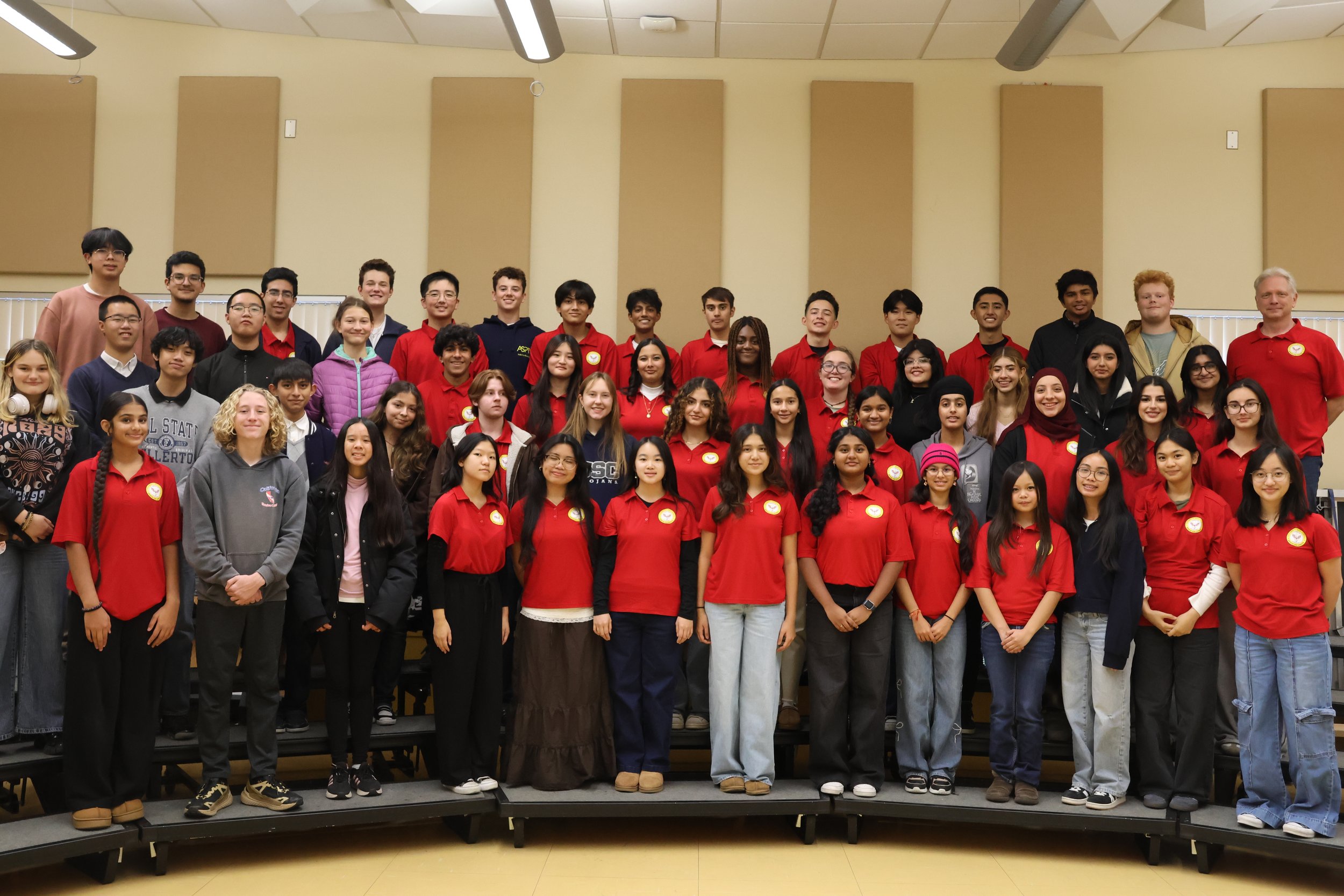 A large group of students and two adults gathered on a tiered platform in a classroom or auditorium, smiling for a group photo. Many students are wearing red shirts, some have headphones, and there are diverse ethnicities and ages.