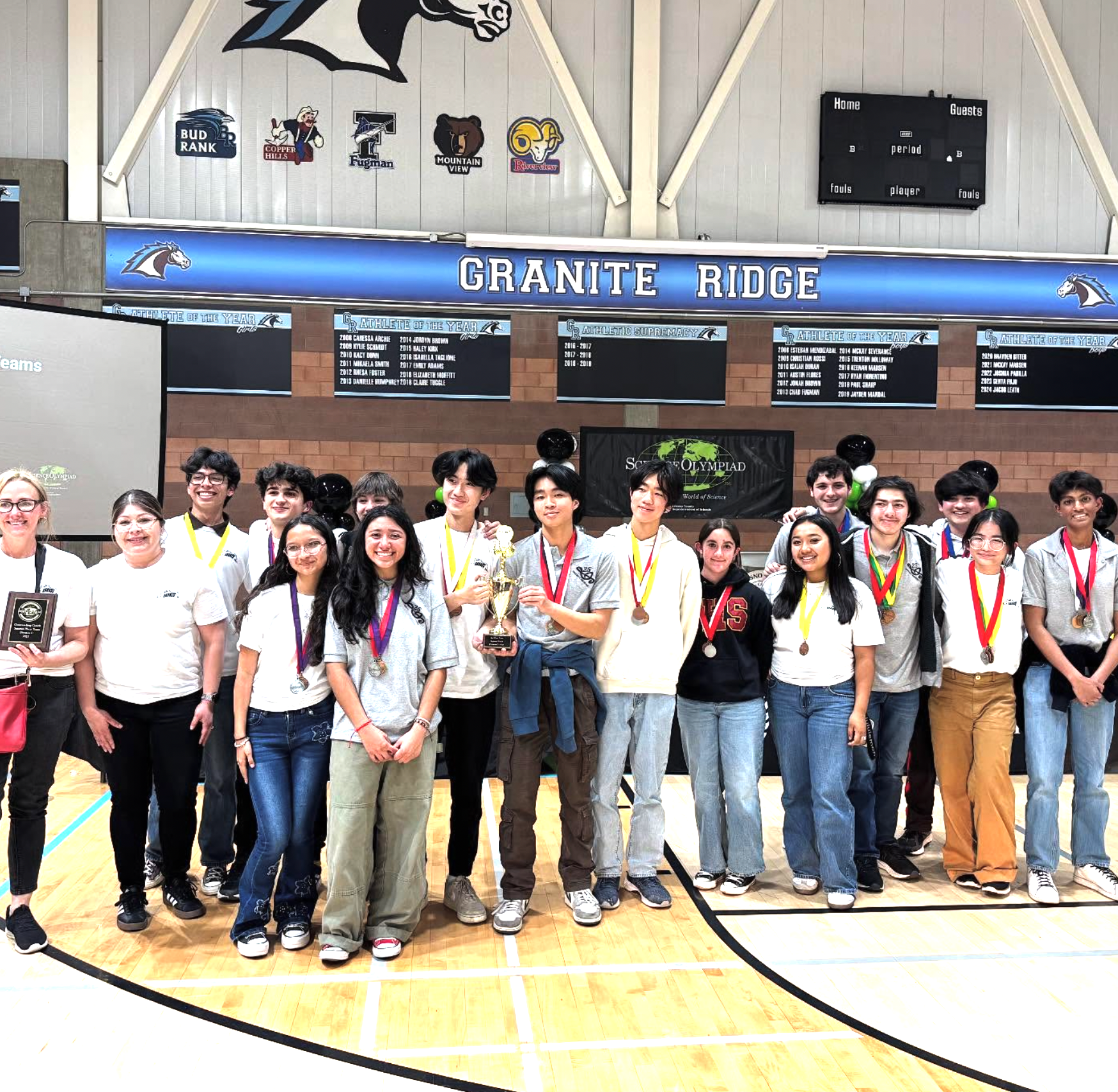 Group of students and teachers in a gymnasium, holding medals and trophies, celebrating an achievement at an academic Olympiad event at Granite Ridge gymnasium.