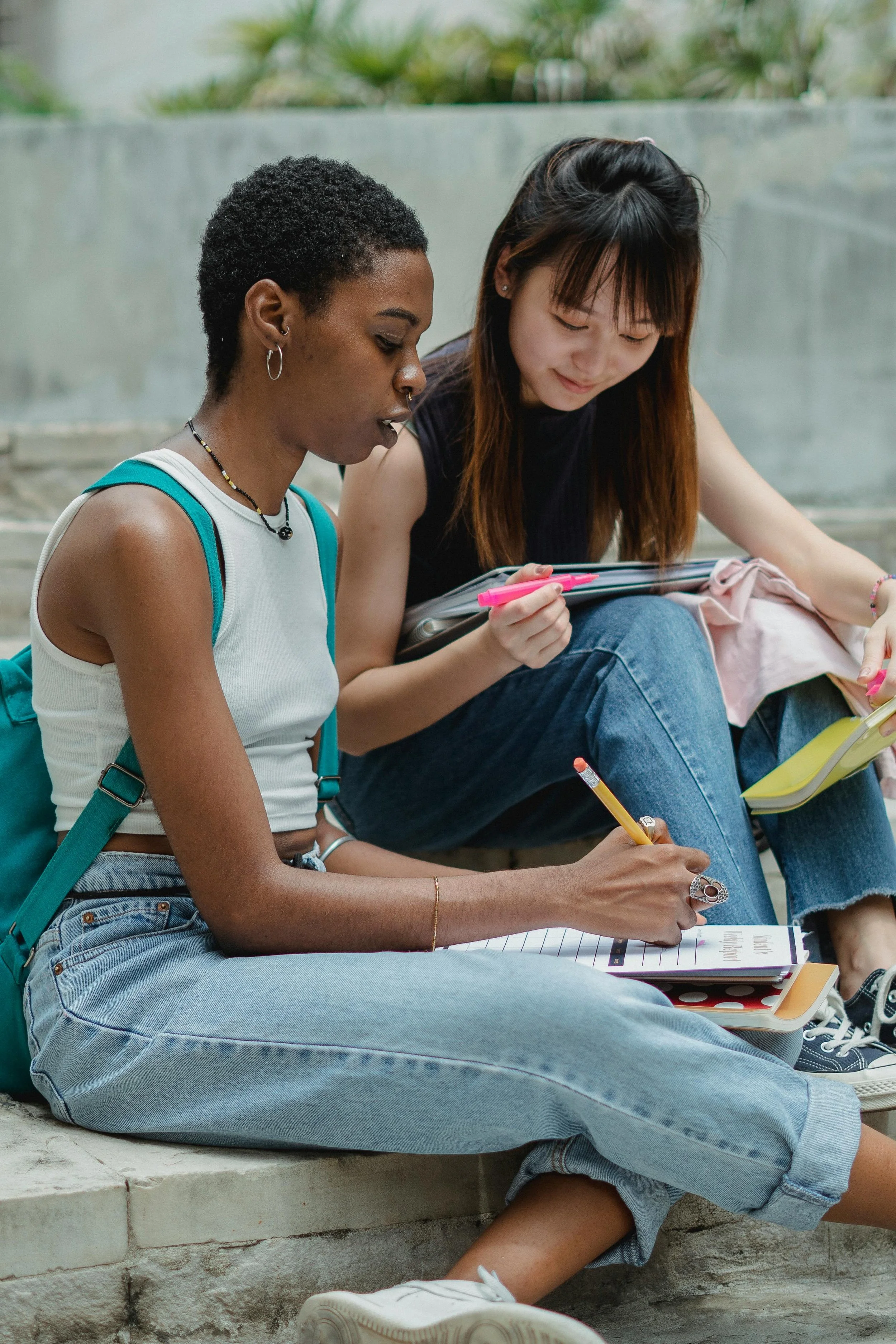 Two young women sit on concrete steps, studying together with notebooks and pens, one with a backpack, outdoors.