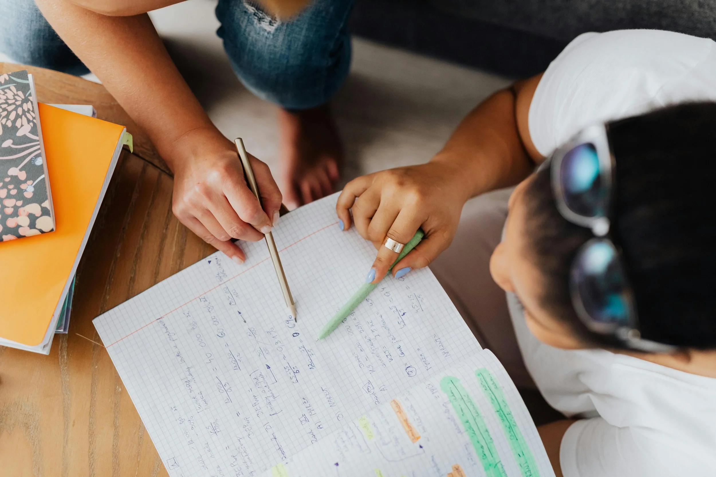 Two people studying together, one writing with a pen, the other pointing at notes on a graph paper notebook on a wooden table.