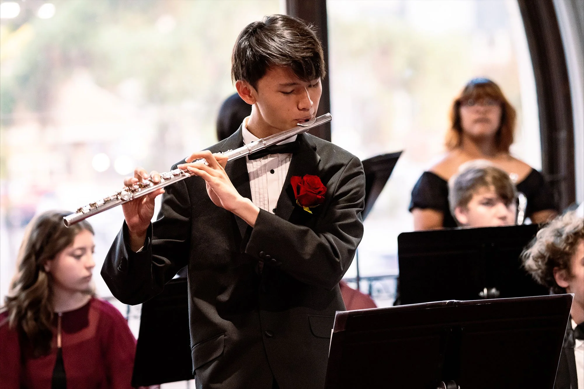 A young man in a black tuxedo with a red rose on his lapel plays a silver flute during a musical performance, with several other musicians and a woman in black behind him.