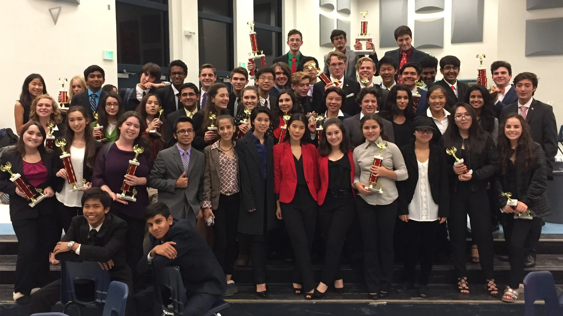 Group of young adults at an award ceremony, holding trophies, smiling, dressed in formal attire, in an indoor venue with large windows and gray walls.