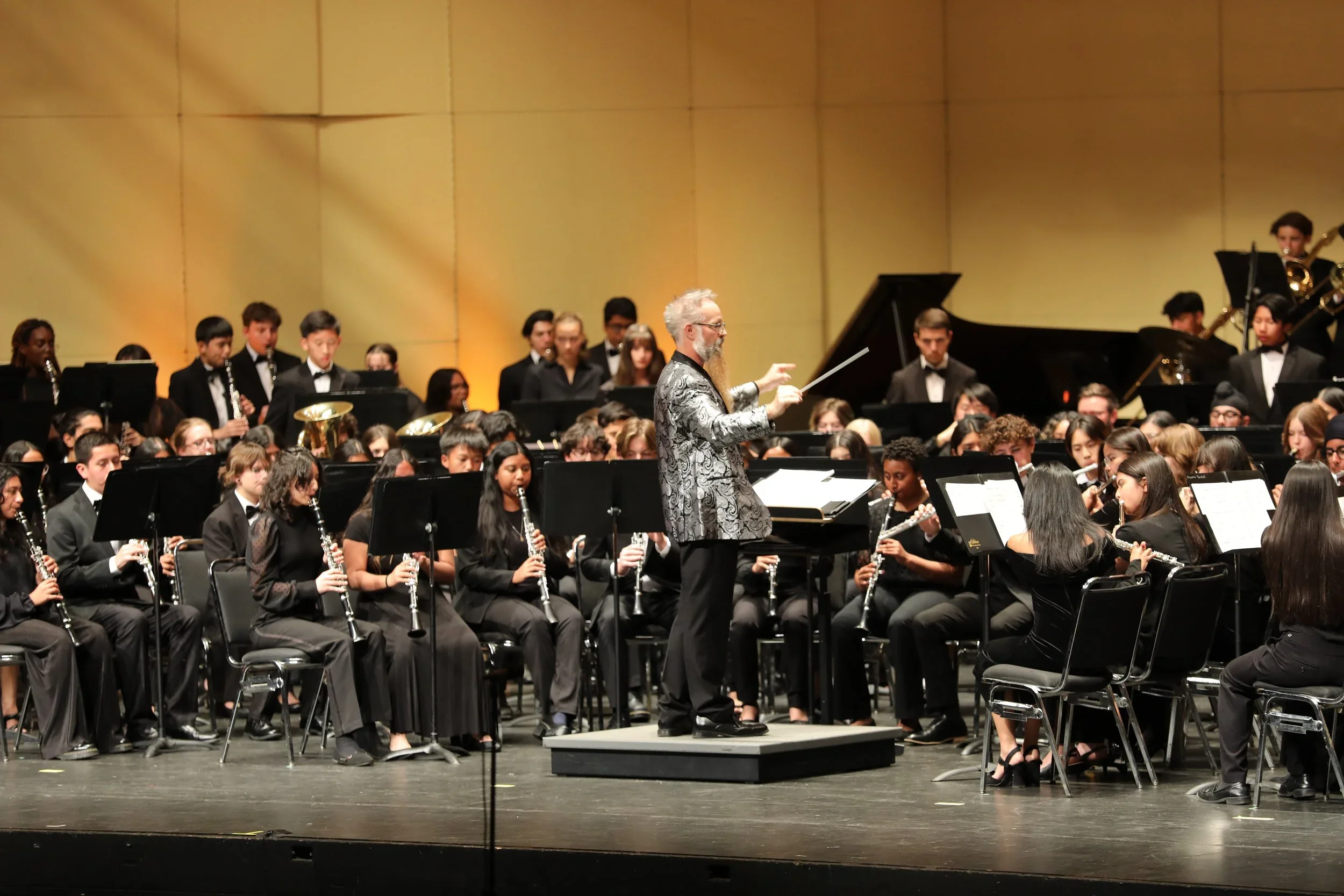 An orchestra performing on stage with a conductor leading the musicians, who are dressed in black formal attire, with a yellow backdrop.