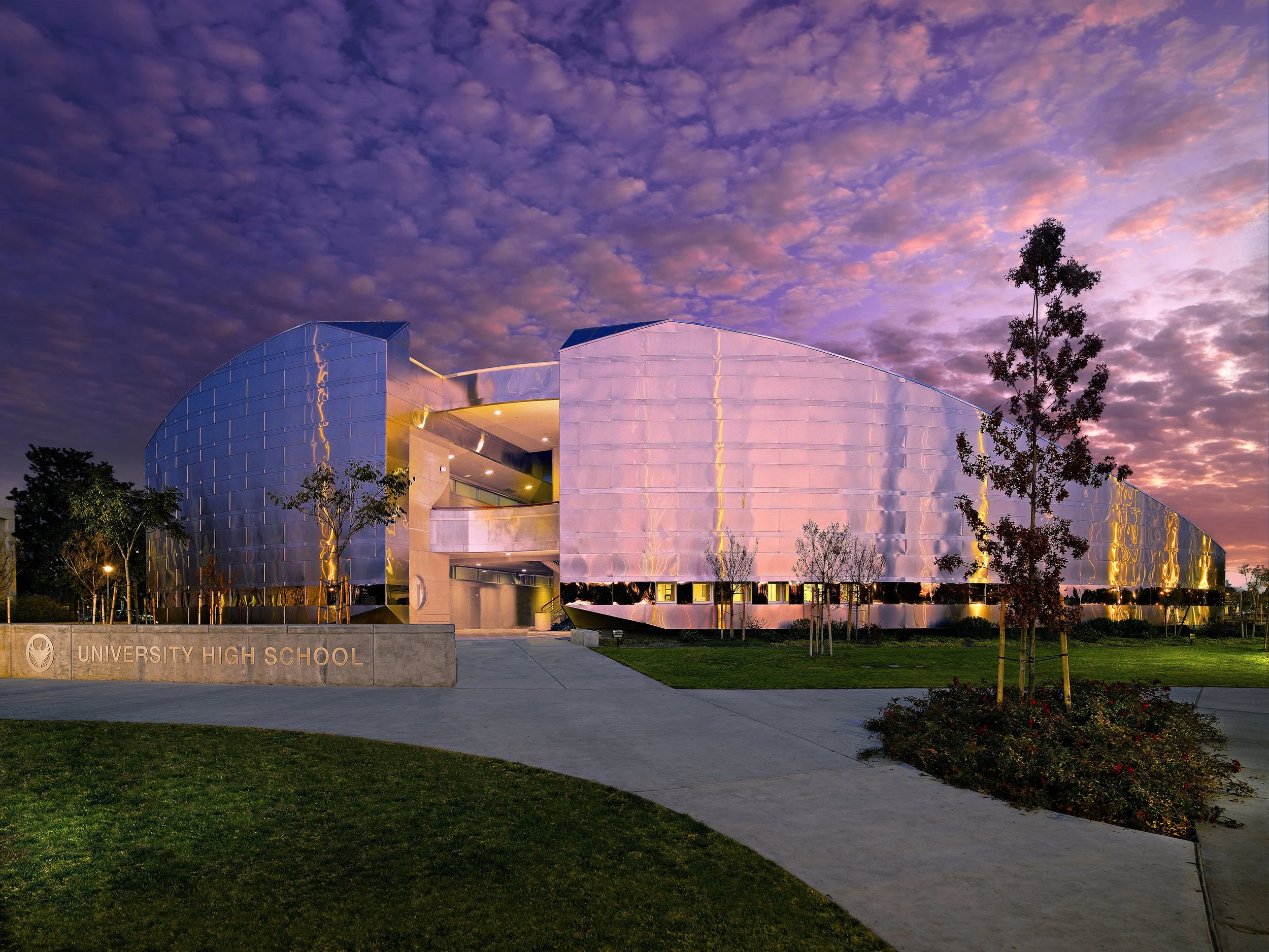 Modern university high school building with reflective metallic facade, trees, and a partly cloudy sky at sunset.