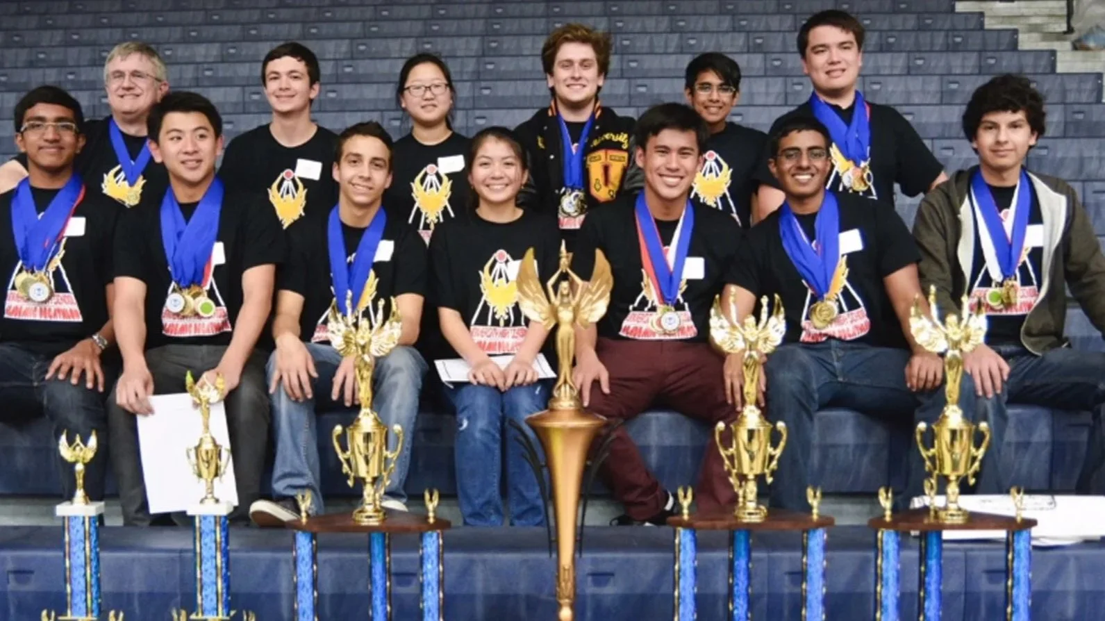 Group of young students and two adults, all wearing medals and black T-shirts with a gold winged emblem, posing with trophies on blue tiered steps in an indoor sports arena.