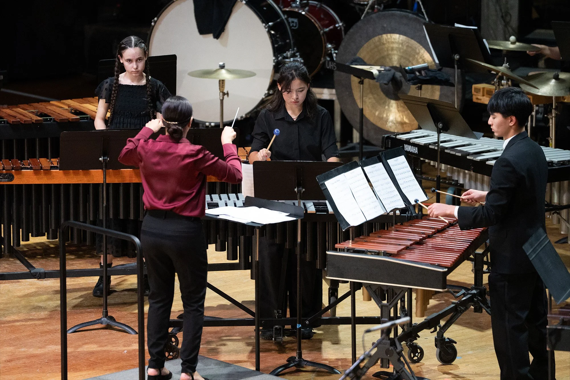 Four musicians performing in a concert hall, playing percussion and xylophones, with sheet music on stands and percussion instruments in the background.