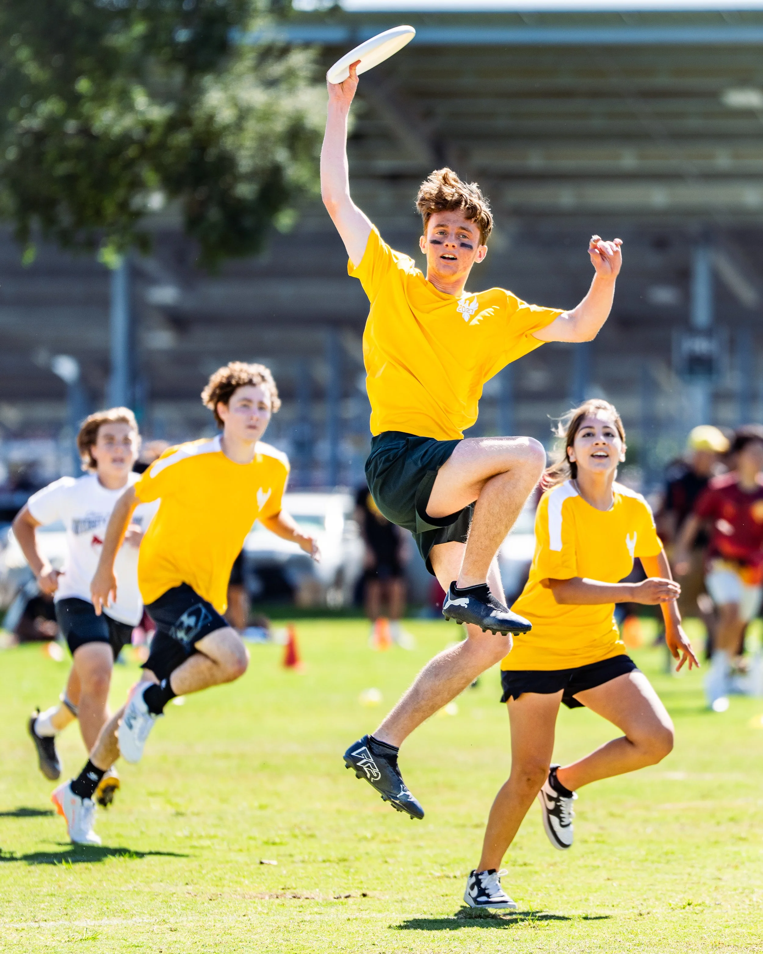 A group of kids playing ultimate frisbee outdoors on a sunny day, with one boy jumping to catch the frisbee while others chase behind him.