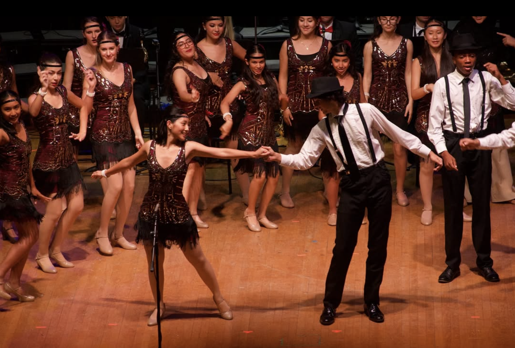 A group of young women and men dressed in vintage-style costumes performing on stage, with the men in white shirts, black suspenders, and hats, and the women in matching dresses, dancing and smiling.