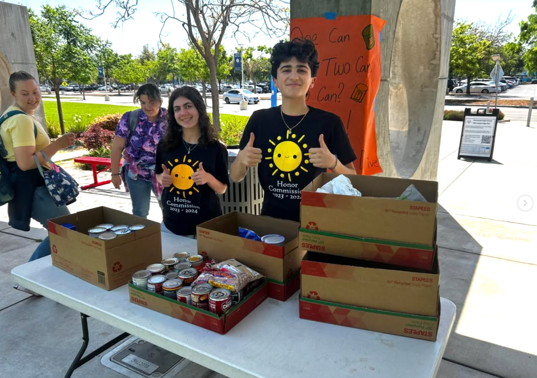 Two students smiling and giving thumbs up at a box of canned goods and food items during a food drive event outside under a shelter. Two other students are in the background, near a sign and a parking lot.