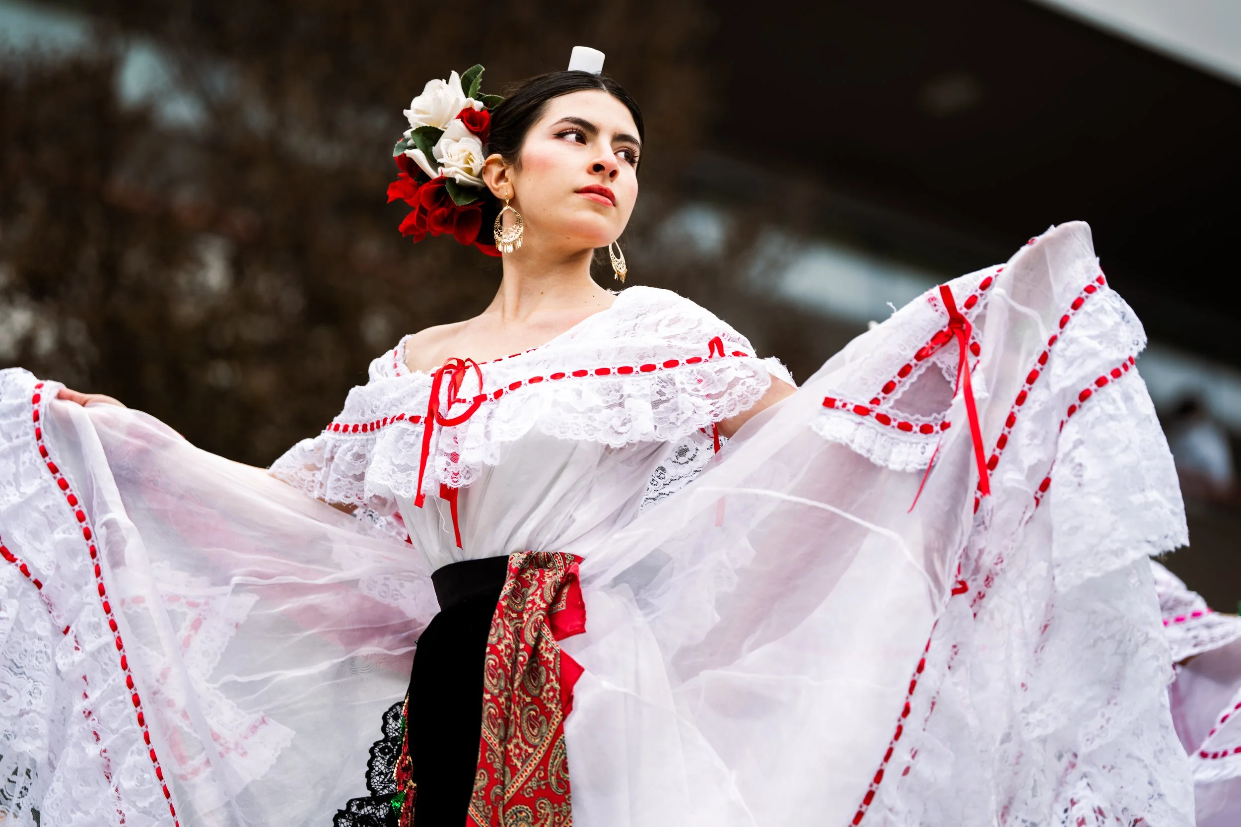 A woman dressed in traditional Mexican clothing performs a dance outdoors. She wears a white dress with red accents and floral headpiece, with a focused expression.