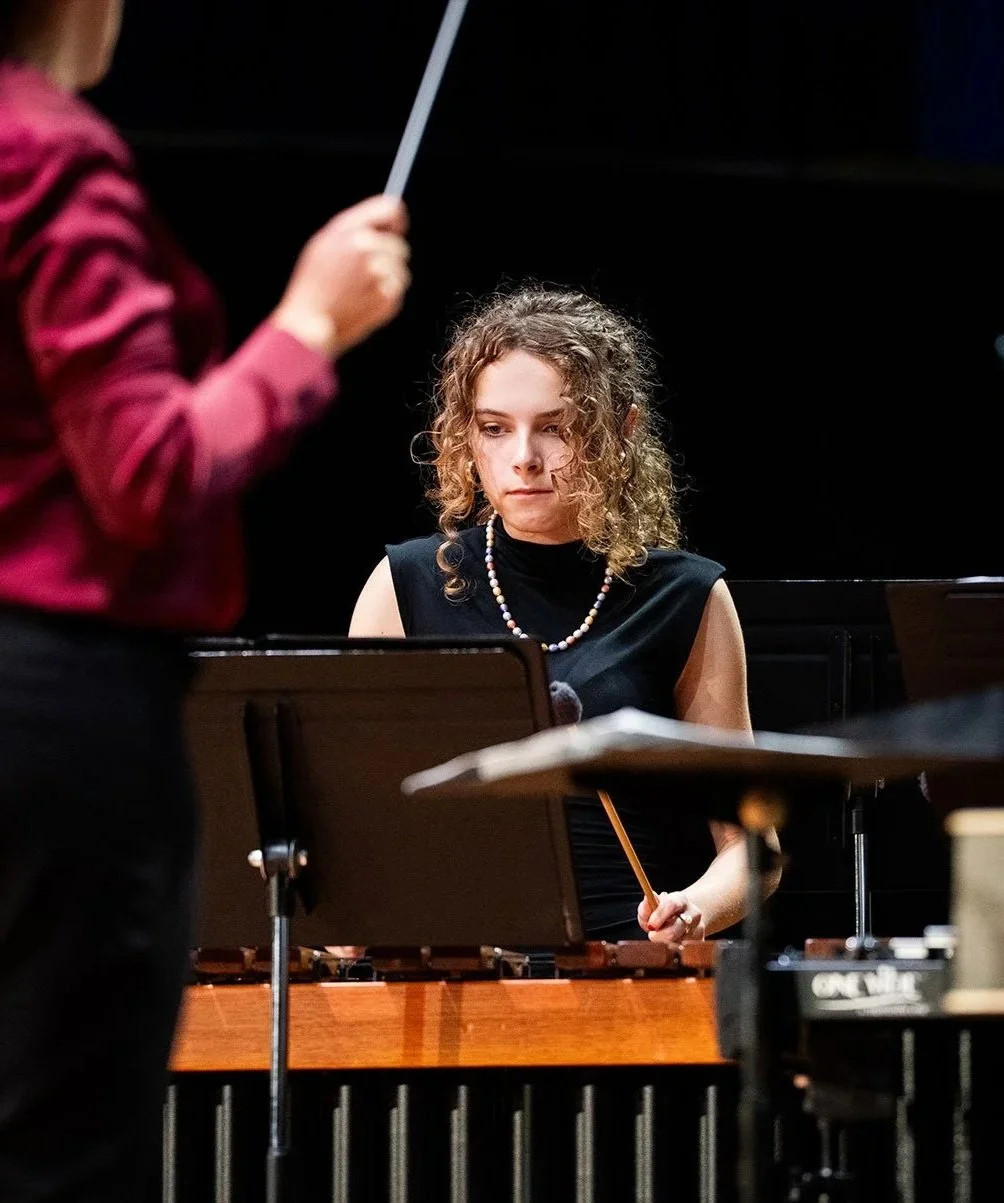 A young woman with curly hair wearing a black dress and a pearl necklace, playing a marimba during a music performance.