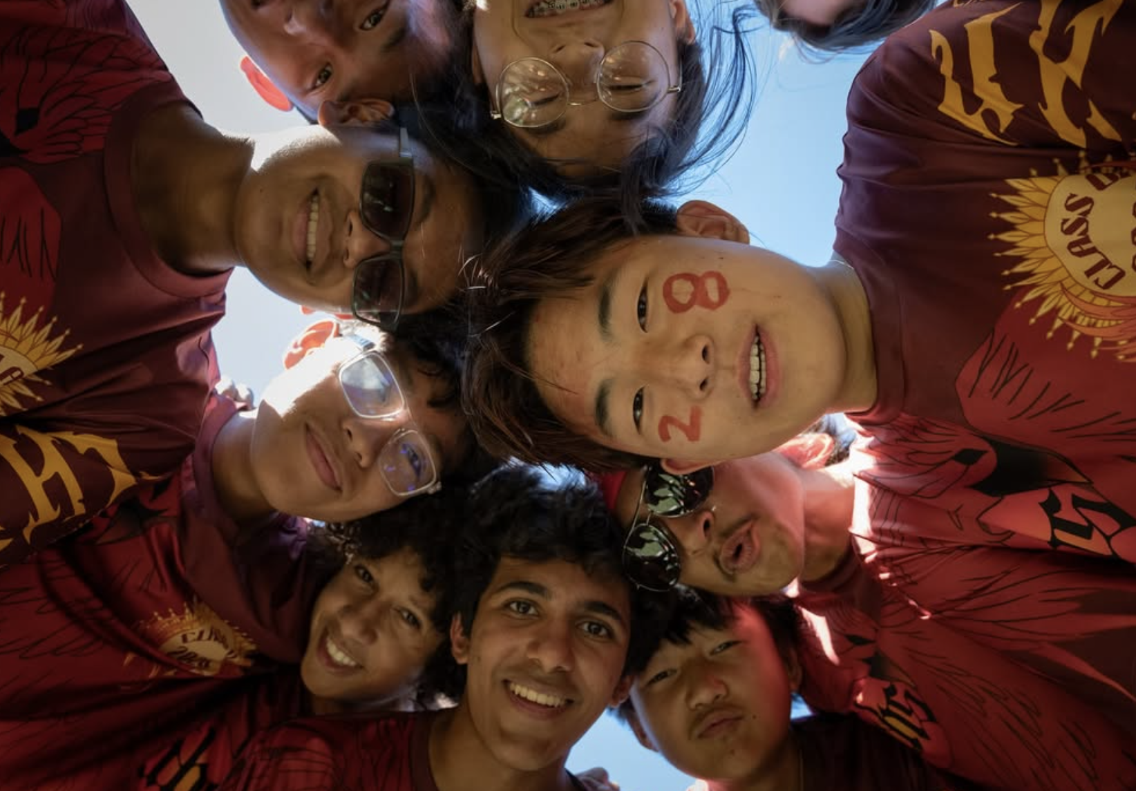 Group of nine young people looking down at camera, forming a circle, outdoors, with blue sky visible, some wearing glasses, and some with face paint, smiling and making playful faces.