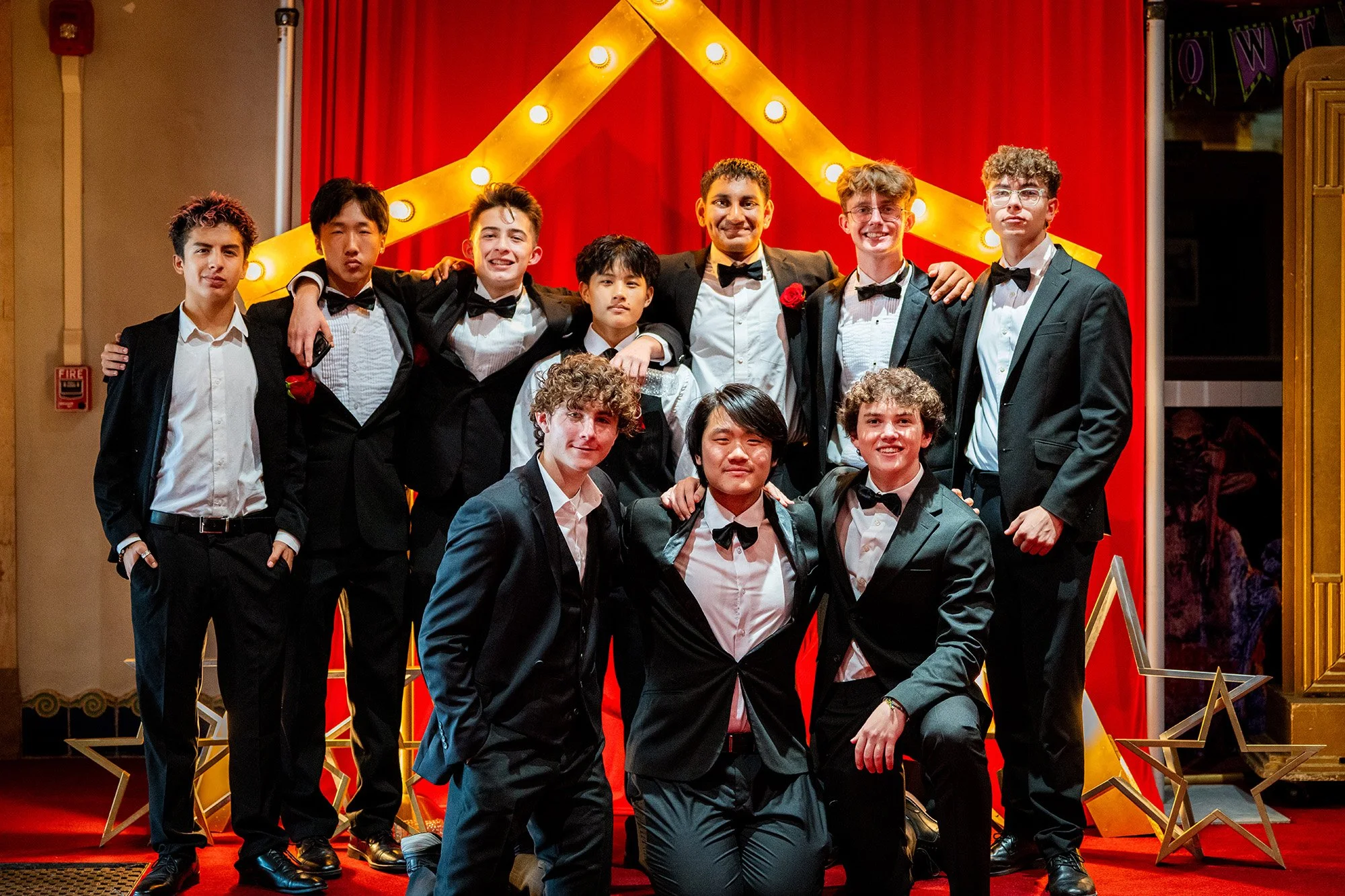 Group of young men dressed in tuxedos at an event, posing in front of a red curtain with a gold star decoration and lights.