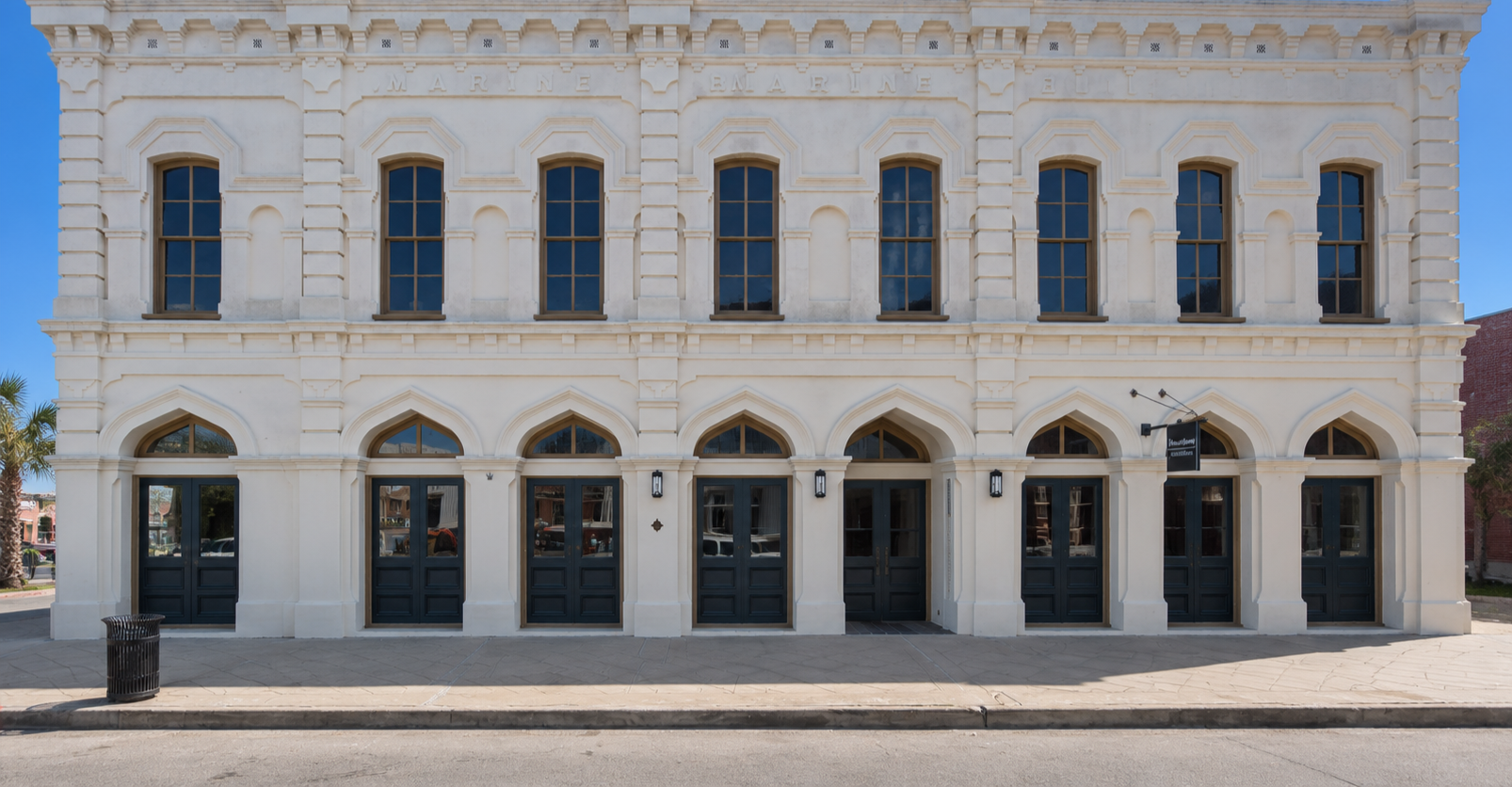 A three-story historic building with white exterior walls and dark windows, featuring arched doorways on the ground floor and rectangular windows on the upper floors, with a sidewalk and street in front.