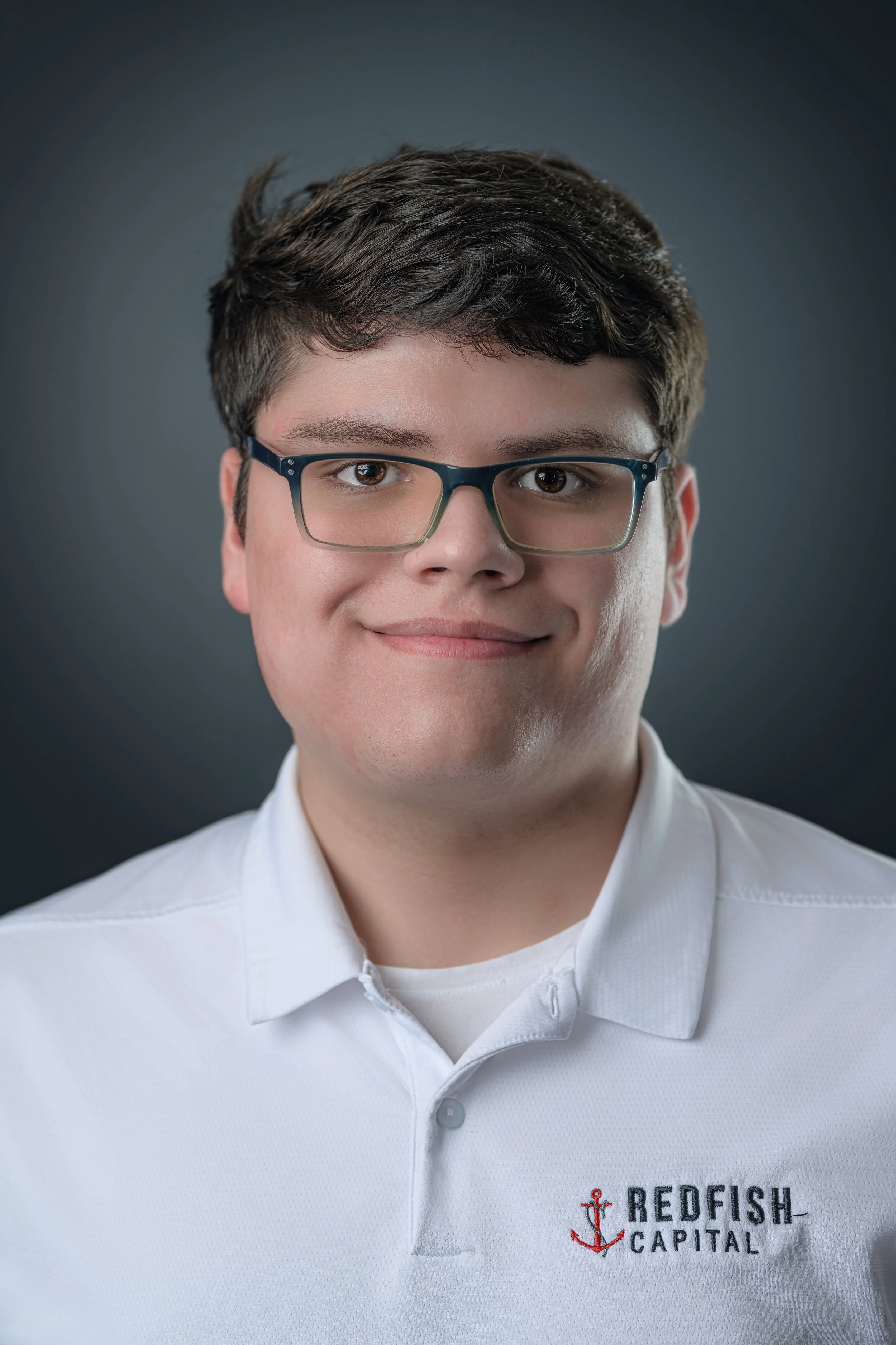 A young man with dark hair, glasses, and a light complexion wearing a white polo shirt with a logo that reads 'Redfish Capital' and features a red anchor.