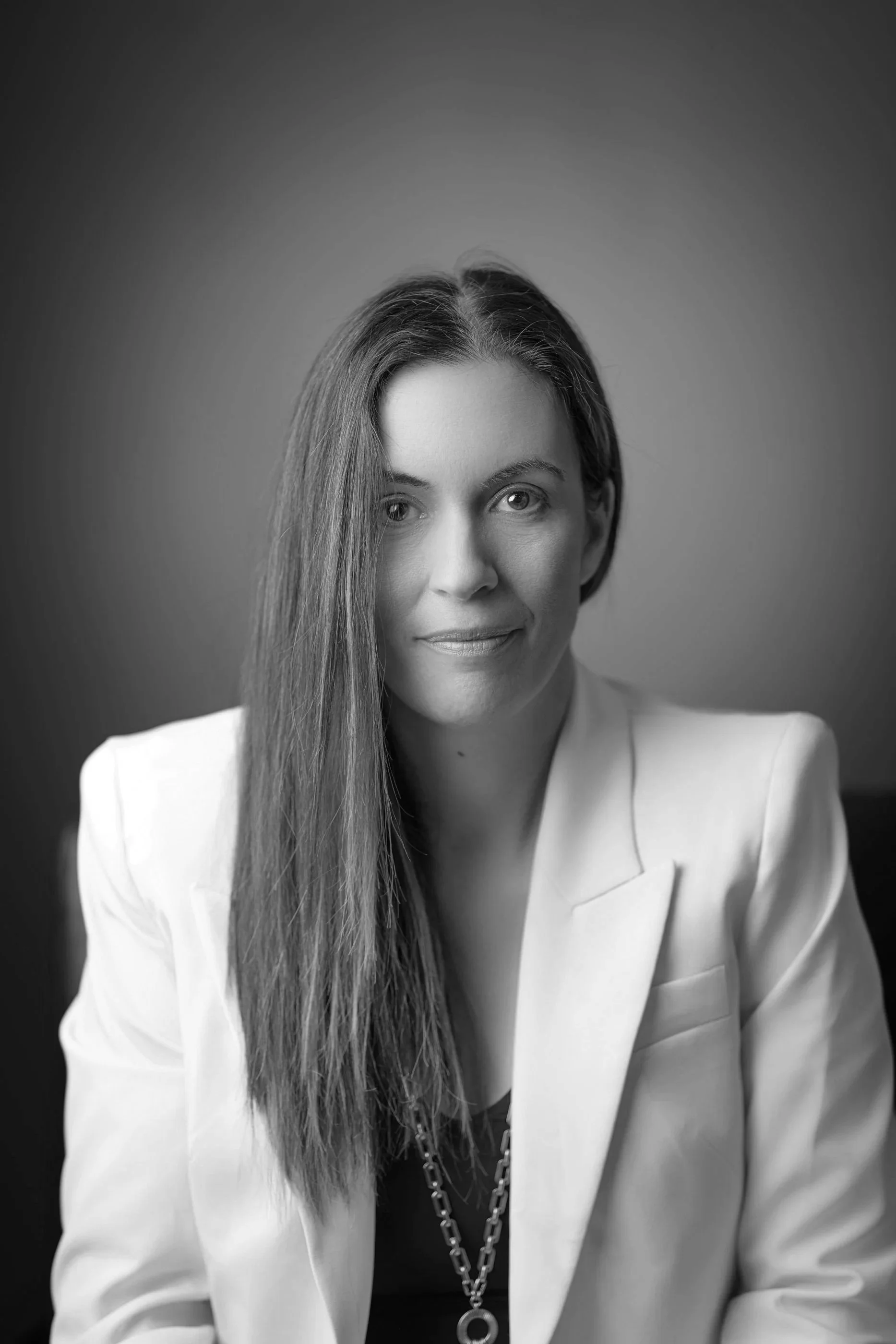 Black and white portrait of a woman with long hair, wearing a white blazer and a necklace, looking at the camera.