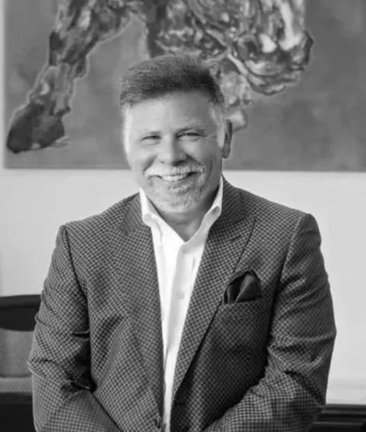 A smiling middle-aged man with a beard and mustache wearing a checked suit jacket and white shirt, sitting in front of an abstract painting.