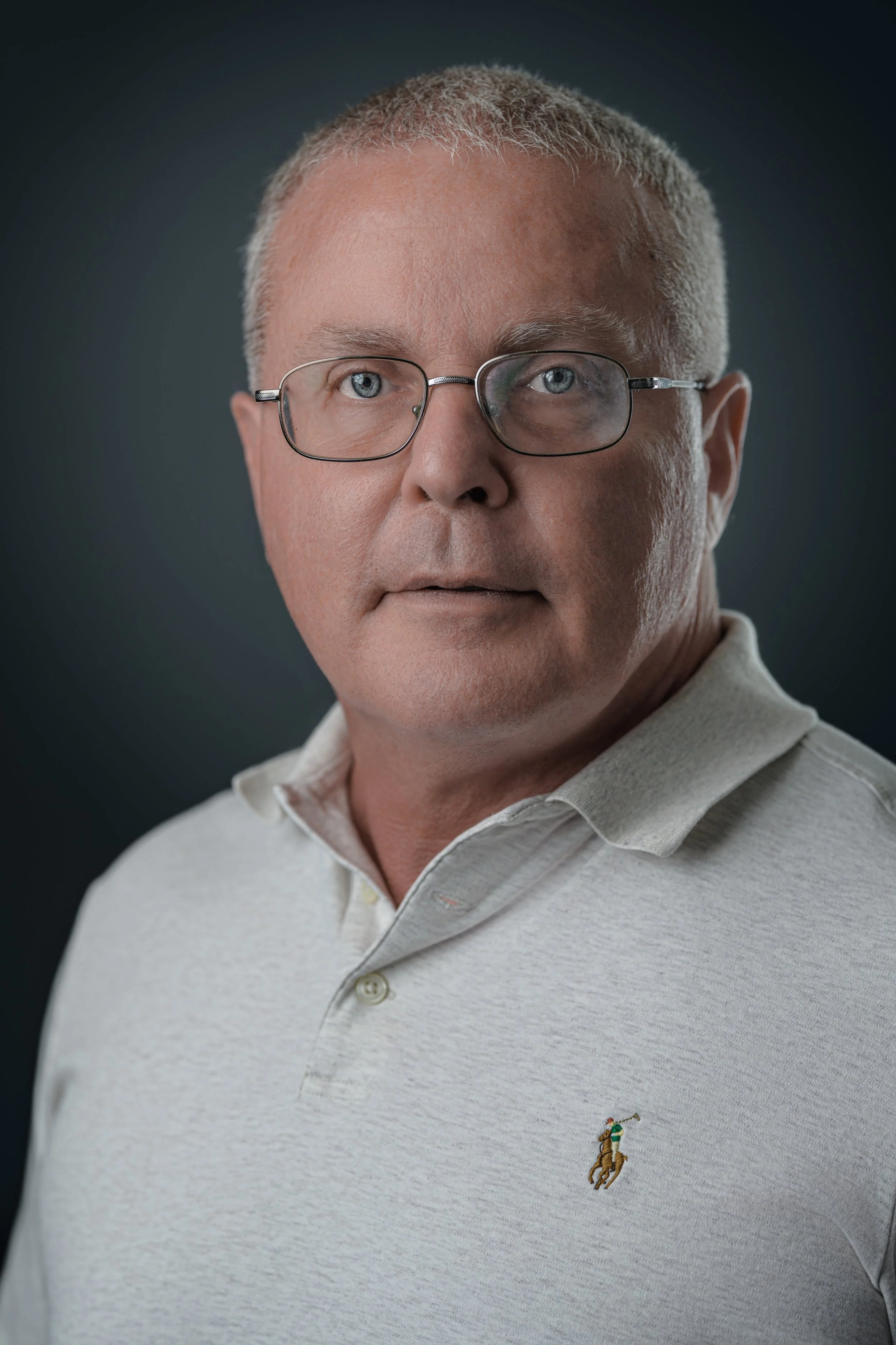 A middle-aged man with short gray hair and glasses, wearing a light-colored collared shirt with a small embroidered polo logo.
