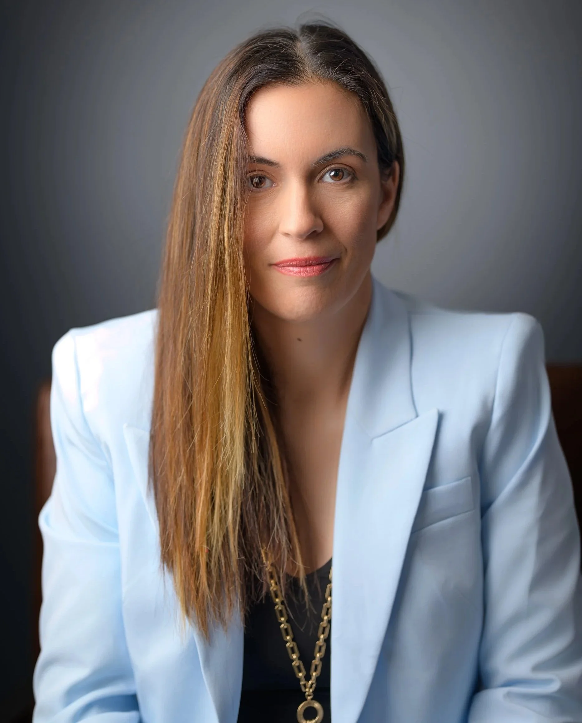 A professional woman with long, straight brown hair, wearing a light blue blazer and a gold chain necklace, looking confidently at the camera.