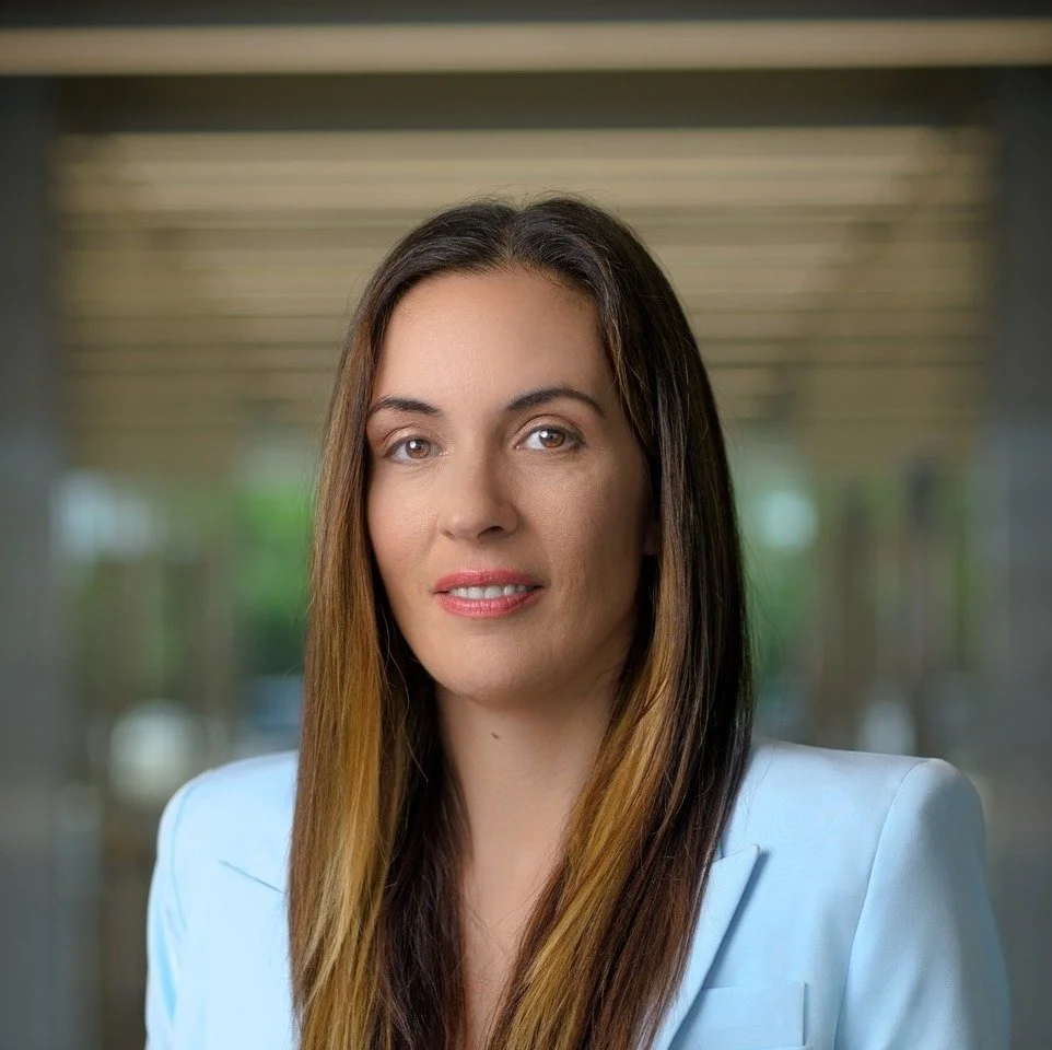 A professional woman with long brown hair and light makeup, wearing a white blazer, standing in an office or modern building with blurred background.