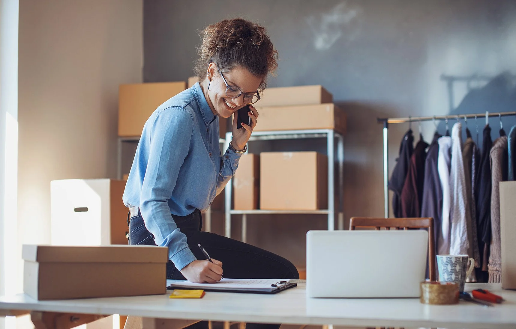 A woman with curly hair and glasses, wearing a denim shirt, is smiling while talking on the phone and writing on a clipboard in a room with boxes and clothing racks.
