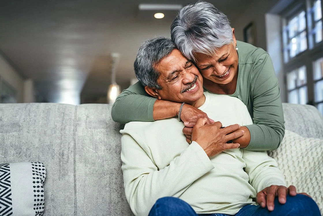 An elderly couple sharing a joyful hug and laughter on a gray couch in a well-lit living room.