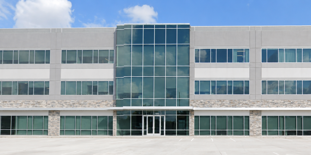 Modern office building with glass exterior, concrete walls, and a parking lot in the foreground under a blue sky with clouds.