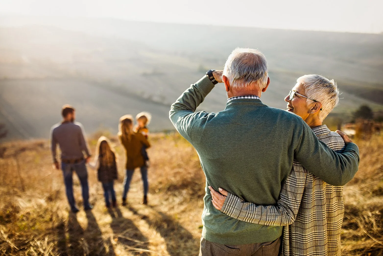 Older couple embracing and smiling in a field during sunset, with a family walking in the background and rolling hills.