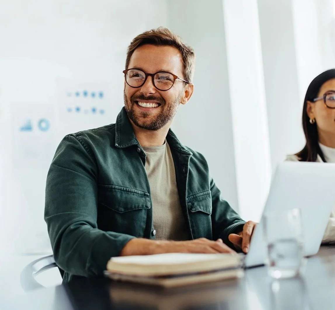 Smiling man wearing glasses and a black jacket working on a laptop in a bright office, with a woman in the background.