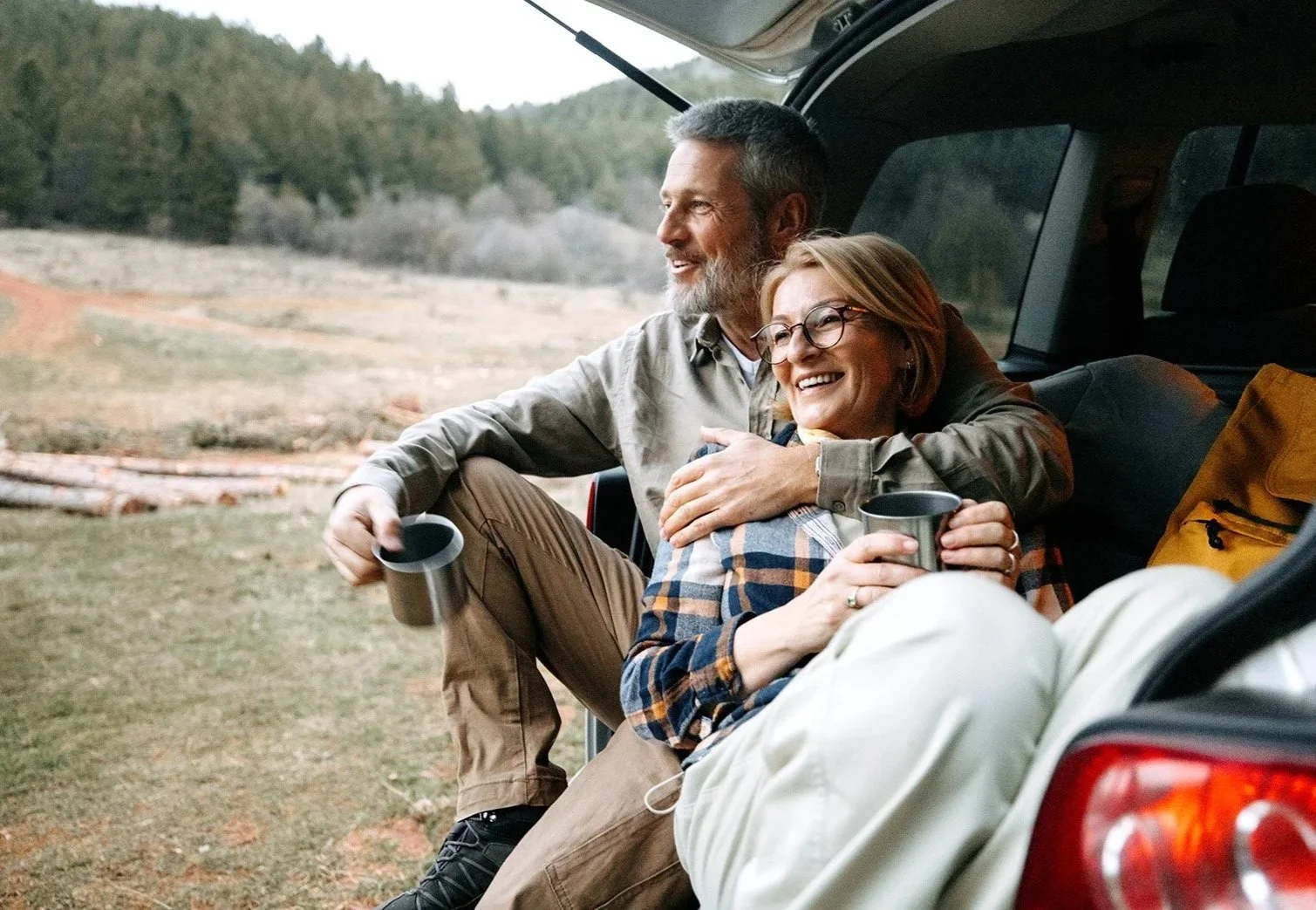 A middle-aged couple sitting at the back of a vehicle, smiling and enjoying a moment outdoors with a scenic mountain landscape in the background. They are holding cups, possibly coffee or tea, and appear happy.