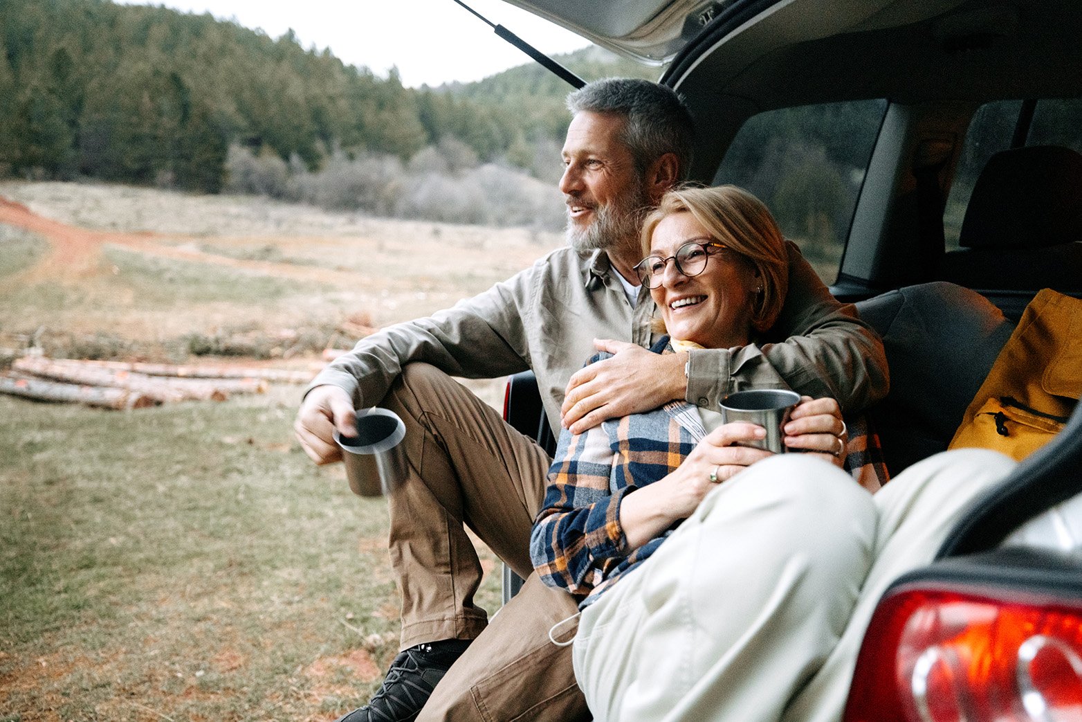 An older couple sitting in an open hatchback of a car in a scenic outdoor setting, smiling, holding mugs, surrounded by nature with a forest in the background.
