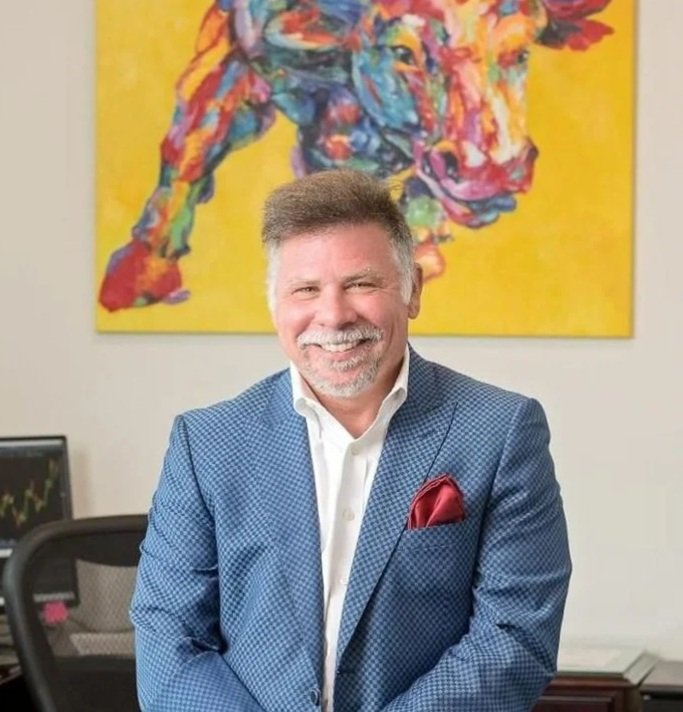 A smiling man with gray hair and a beard wearing a blue checked suit with a white shirt and red pocket square, standing in an office with a colorful bull painting on the wall behind him.