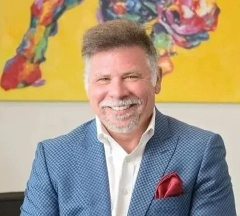 A smiling man with gray hair and a beard, wearing a blue checkered blazer, a white shirt, and a red pocket square, standing in front of a colorful abstract painting.