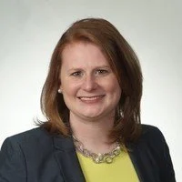 A woman with shoulder-length red hair, wearing a dark blazer and a yellow top, smiling against a light background.