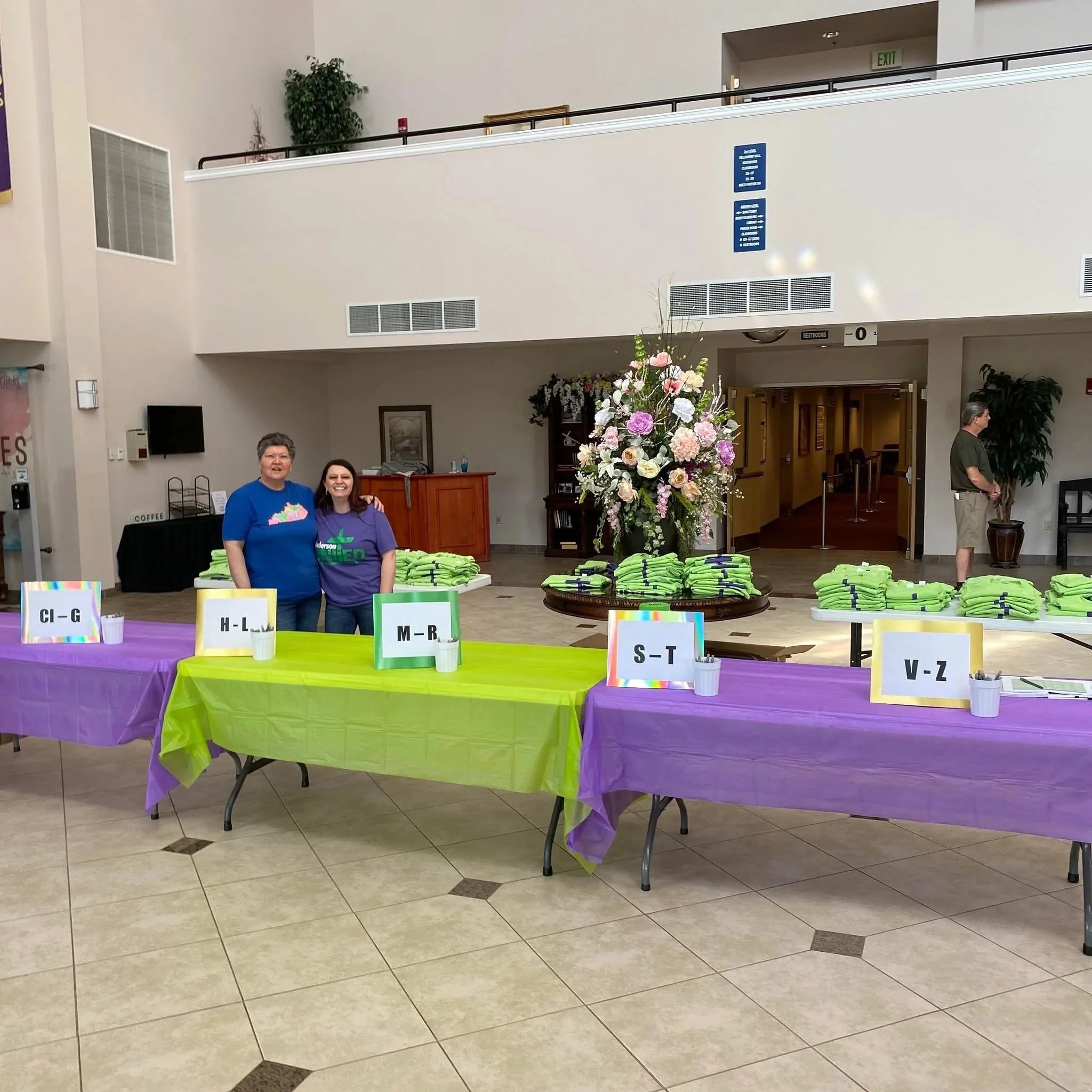 Registration tables with colorful tablecloths and organized stacks of green T-shirts, divided into alphabetic sections. Two women stand behind the tables, smiling, in a spacious indoor event space with decorative flowers and a person in the background.