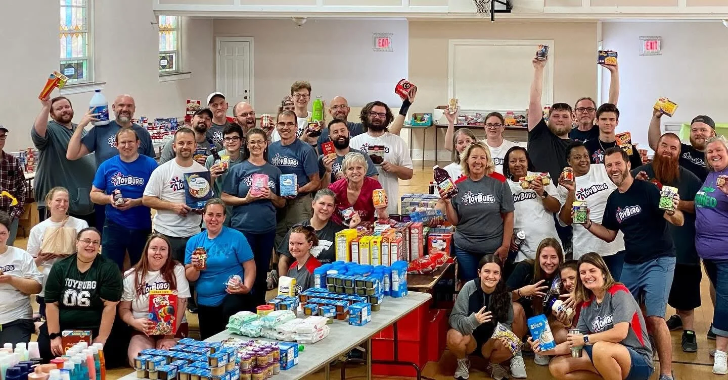 Group of people inside a community center or hall, holding food items and toiletries, standing behind tables with supplies, smiling for a group photo during a charity event or donation drive.