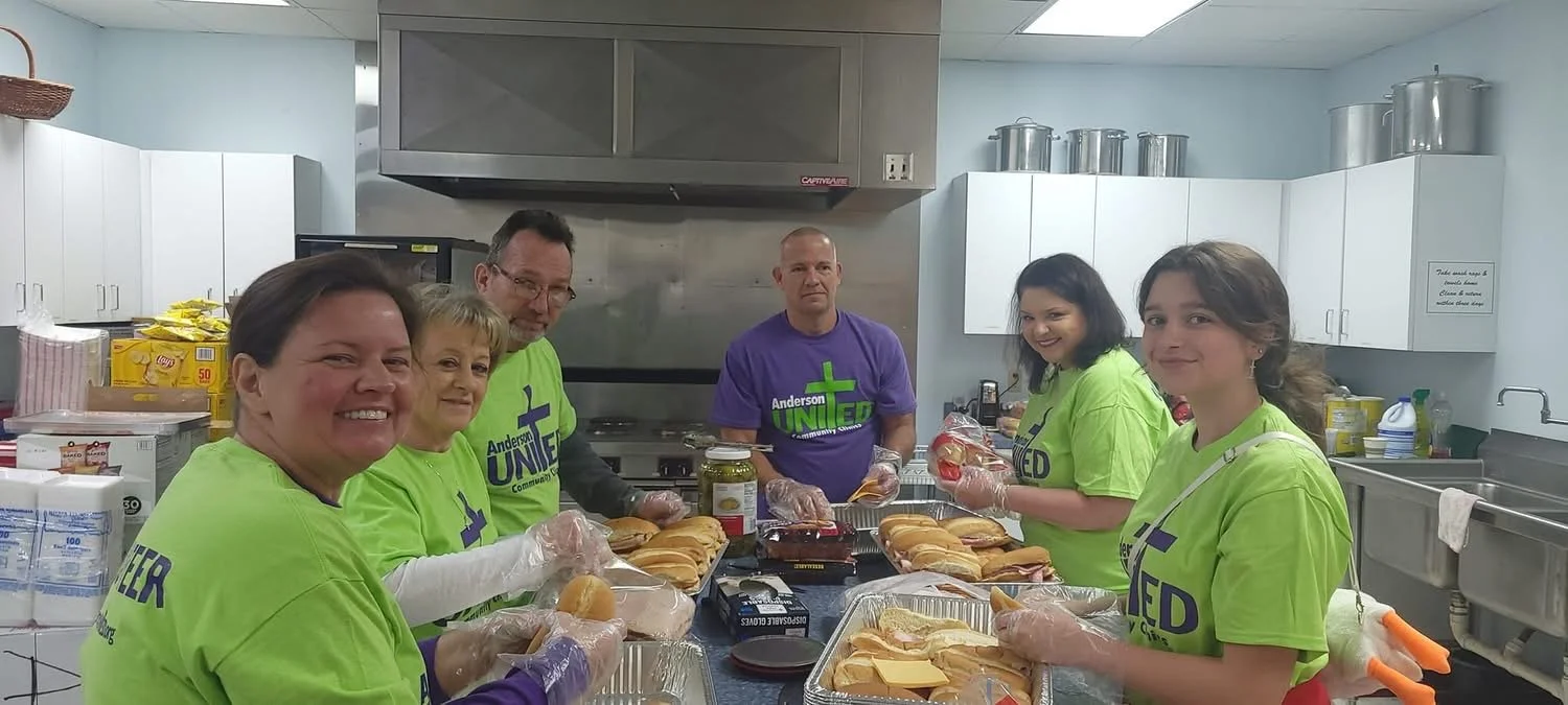 Group of people preparing sandwiches in a kitchen. They are wearing green shirts with a community organization logo and disposable gloves.