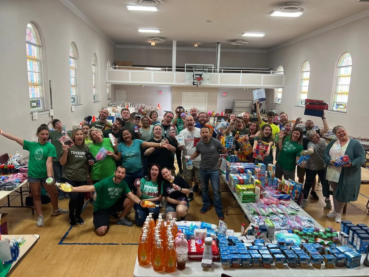 A large group of smiling people gathered inside a gymnasium with tables filled with various donated items like food and toiletries, celebrating a successful charity event or volunteer activity.