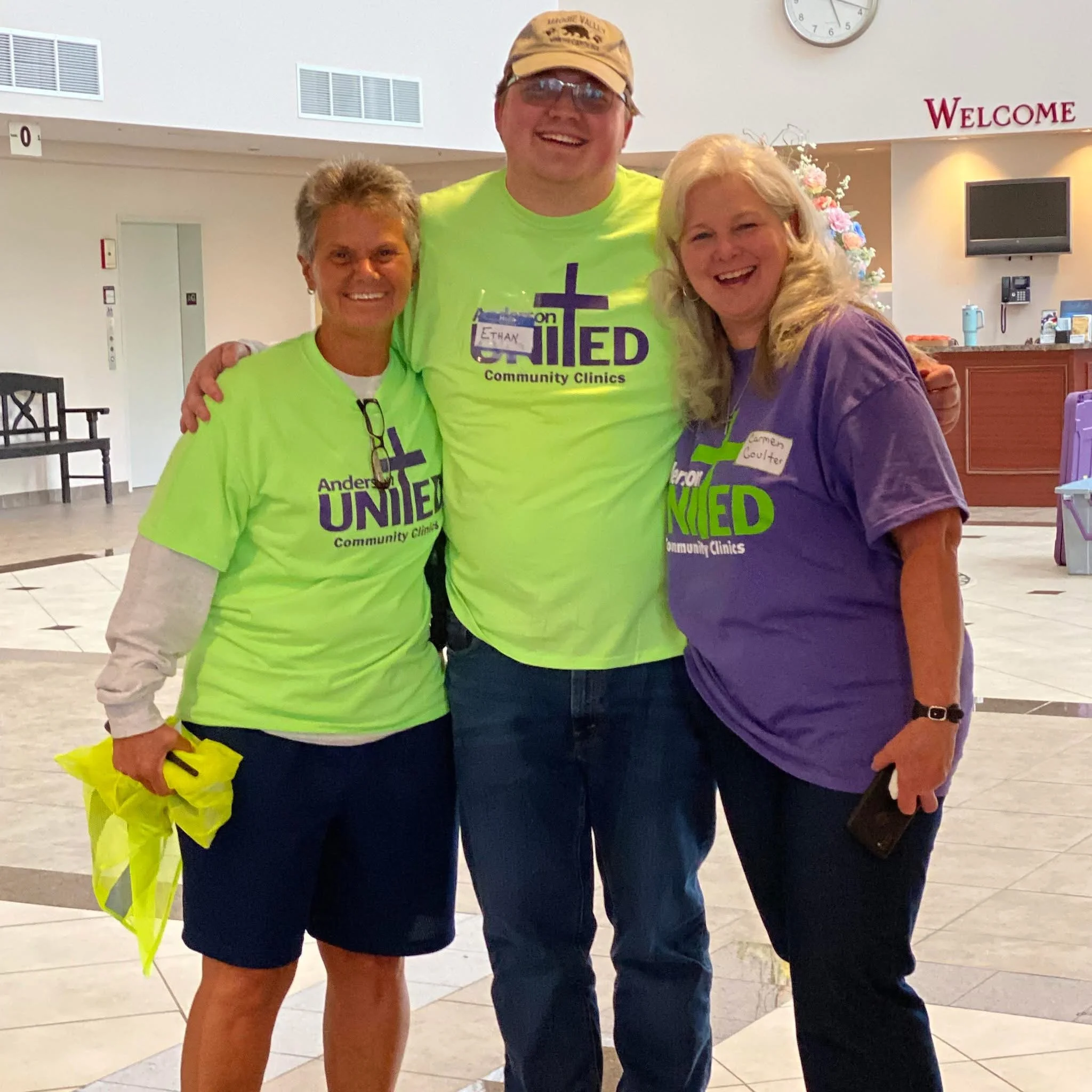 Three people standing together inside a building, smiling, wearing bright green and purple shirts with 'United Community Clinics' logo, and posing for the camera.
