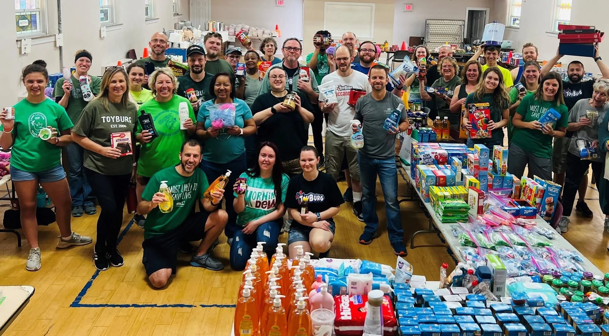 Group of people gathered inside a community center, holding various donated food and household items, with tables displaying more supplies and snacks.
