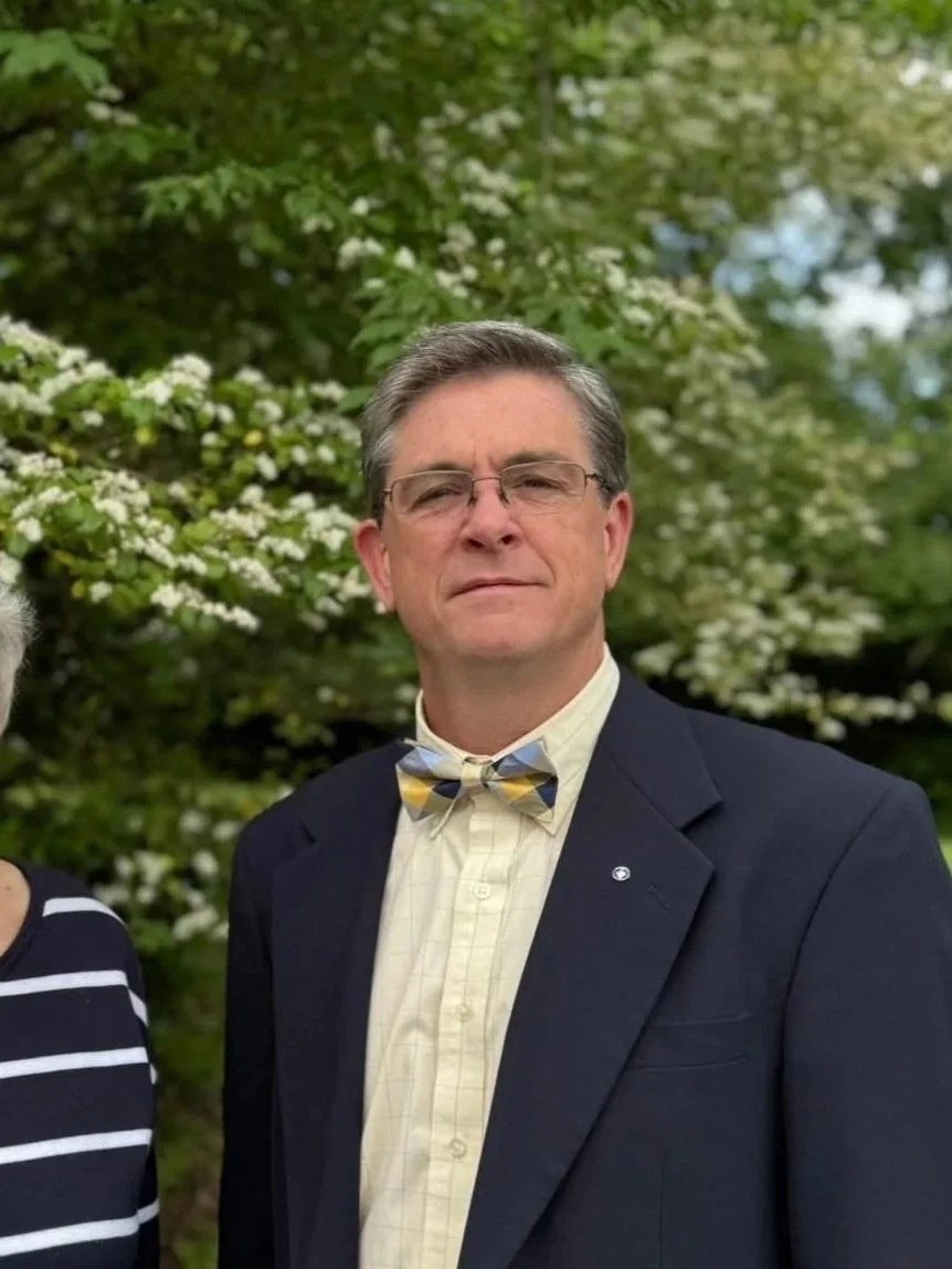A man wearing glasses, a dark blazer, a yellow checkered shirt, and a multicolored bowtie standing outdoors with green foliage and white flowering bushes in the background.