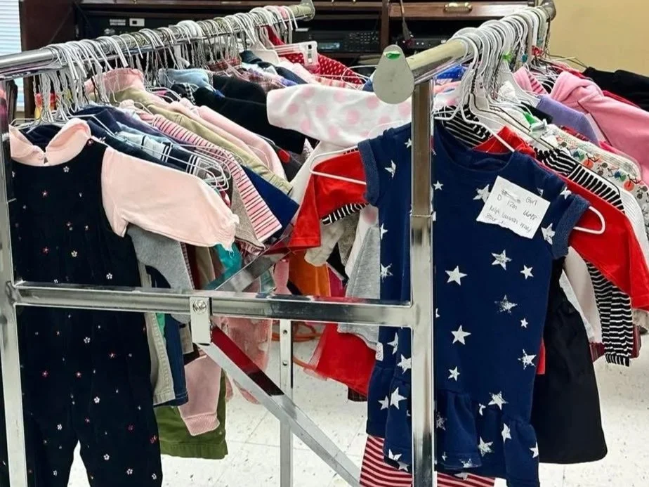 Clothing on a metal rack in a thrift or donation store, including children's dresses, shirts, and pajamas with patriotic and colorful patterns.